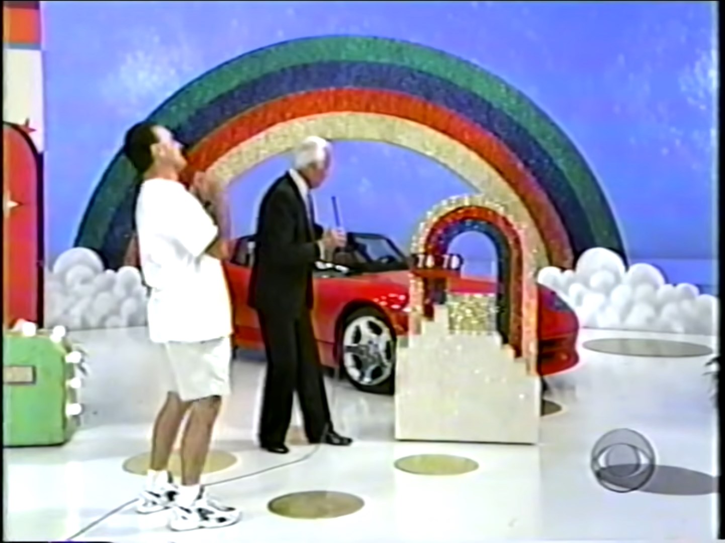 A contestant celebrates with a host near a red car and a colorful rainbow display on a game show set.