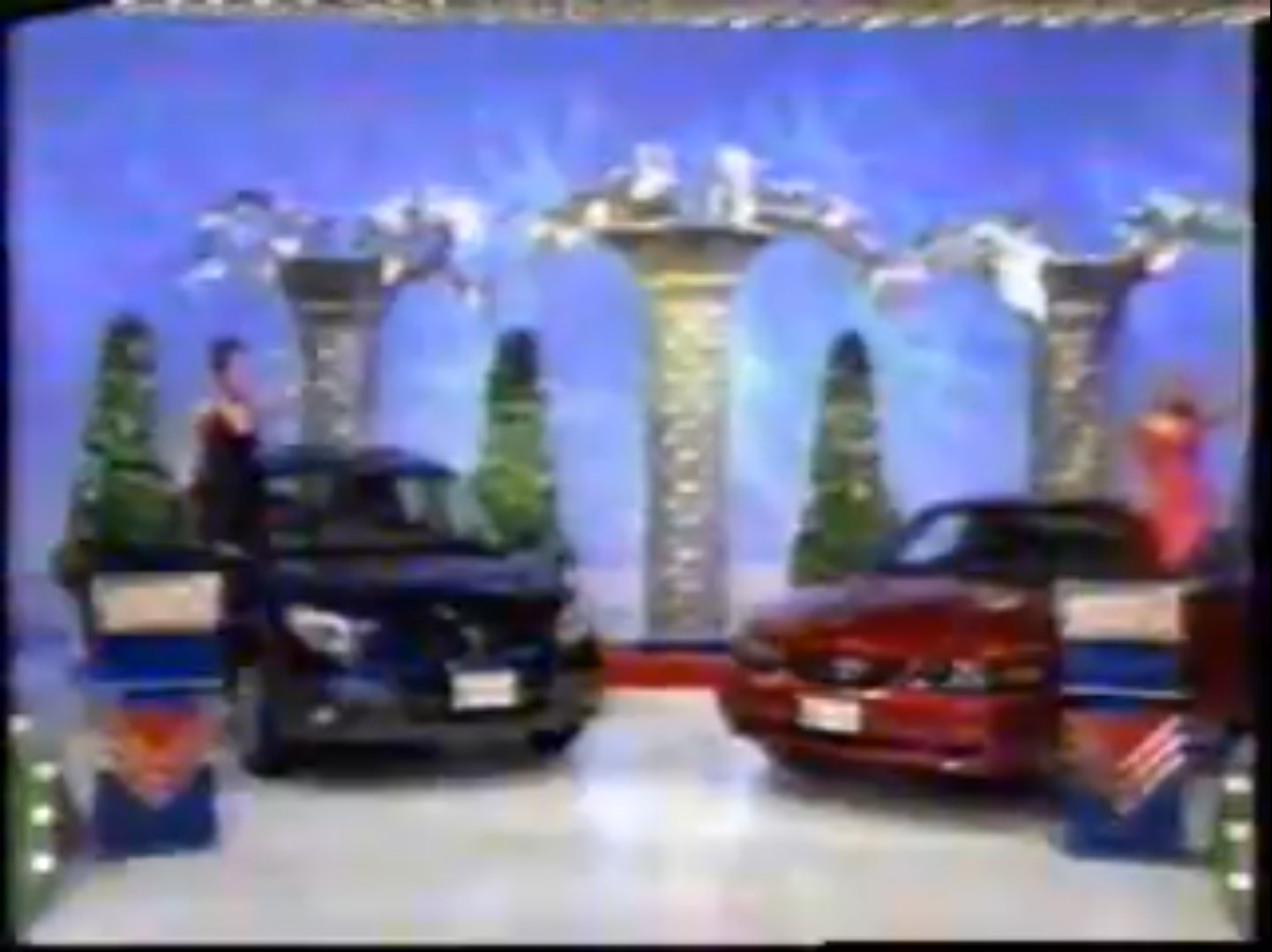Two cars displayed on a game show set, with a contestant and audience visible, set against a festive backdrop.