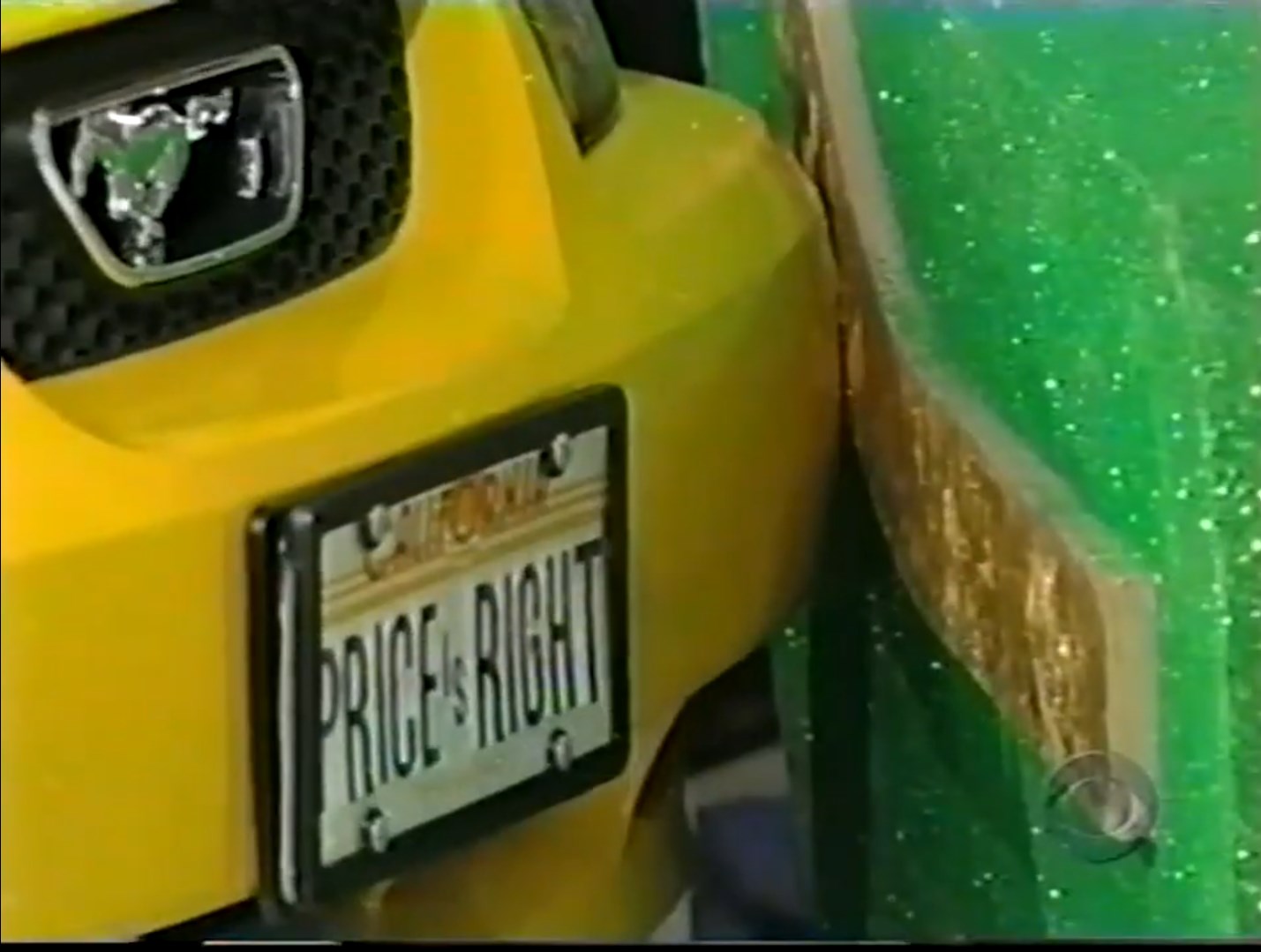 Close-up of a yellow car with a 'Price is Right' license plate, partially crashed into a green sparkly backdrop.