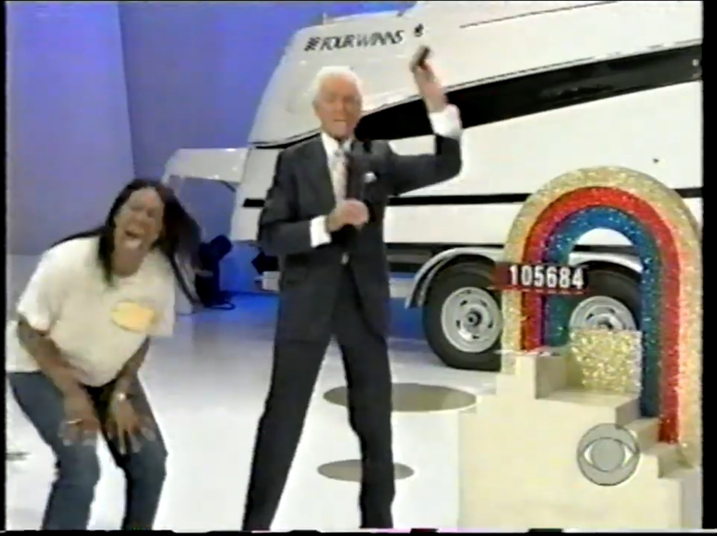 A cheerful contestant laughs and bends down in excitement while the host, dressed in a suit, gestures with a microphone in hand against a backdrop featuring a large RV and a colorful rainbow display.