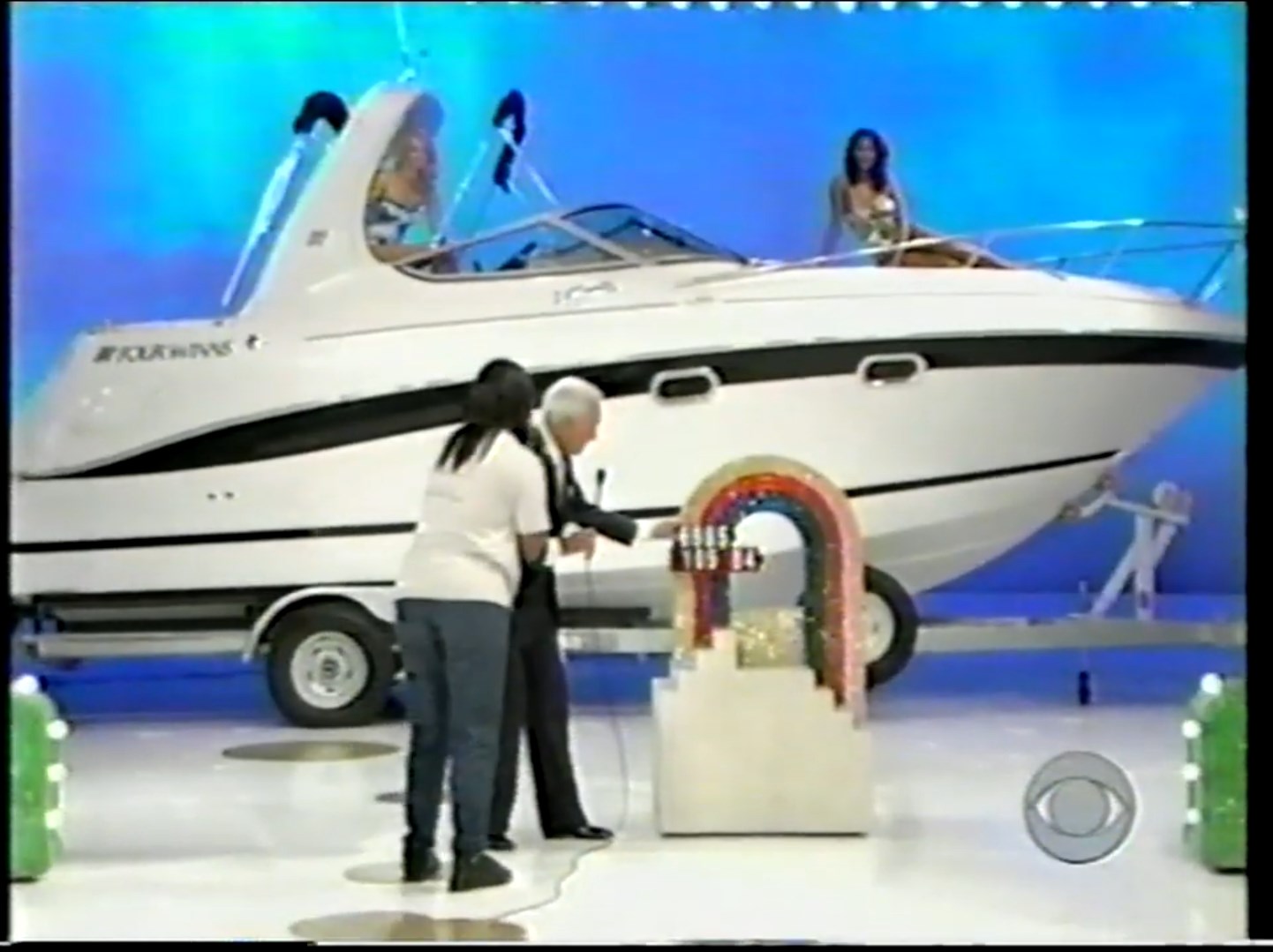 Contestant interacting with the host beside a Four Winns cabin cruiser on the set of The Price Is Right, with a colorful prize display in the foreground.