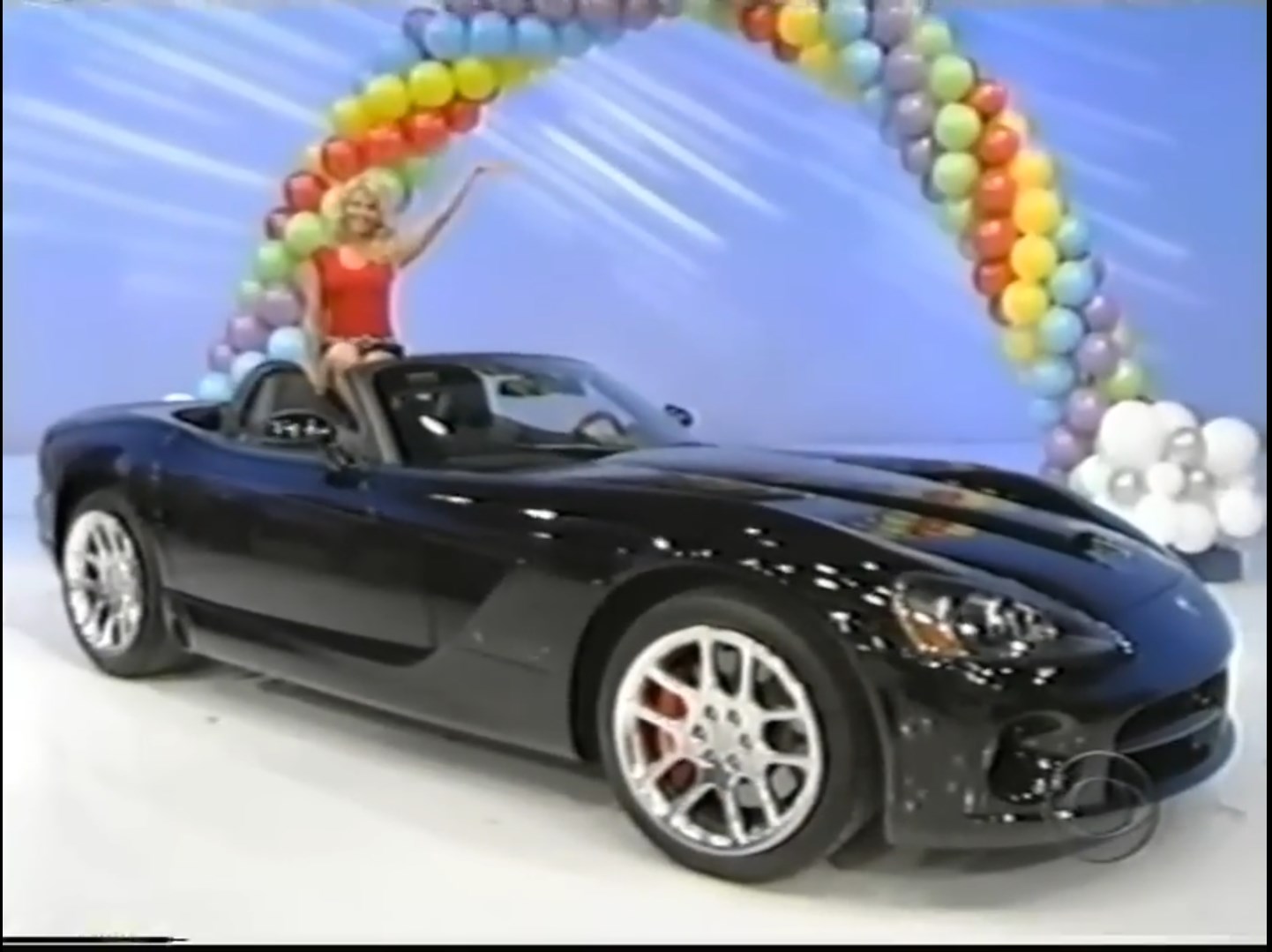 A black sports car with prominent wheels showcased under a colorful balloon arch, and a woman in a red outfit sitting atop the car, smiling and waving.