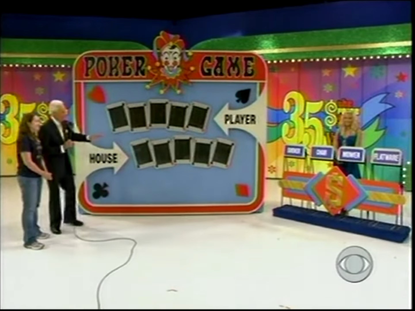 A scene from the 'Poker Game' segment on a game show, featuring a host pointing towards a board with cards representing the house and player. Contestants stand beside the host, with colorful background decorations visible.