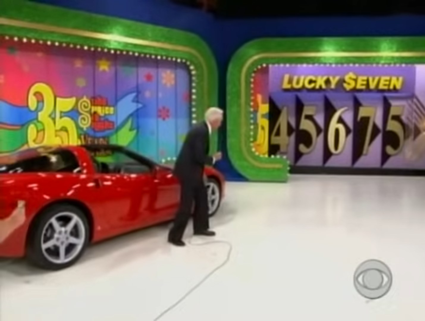 Bob Barker stands near a red Corvette sports car on stage on his final episode with colorful graphics and numbers displayed behind him, indicating the game 'Lucky Seven'.