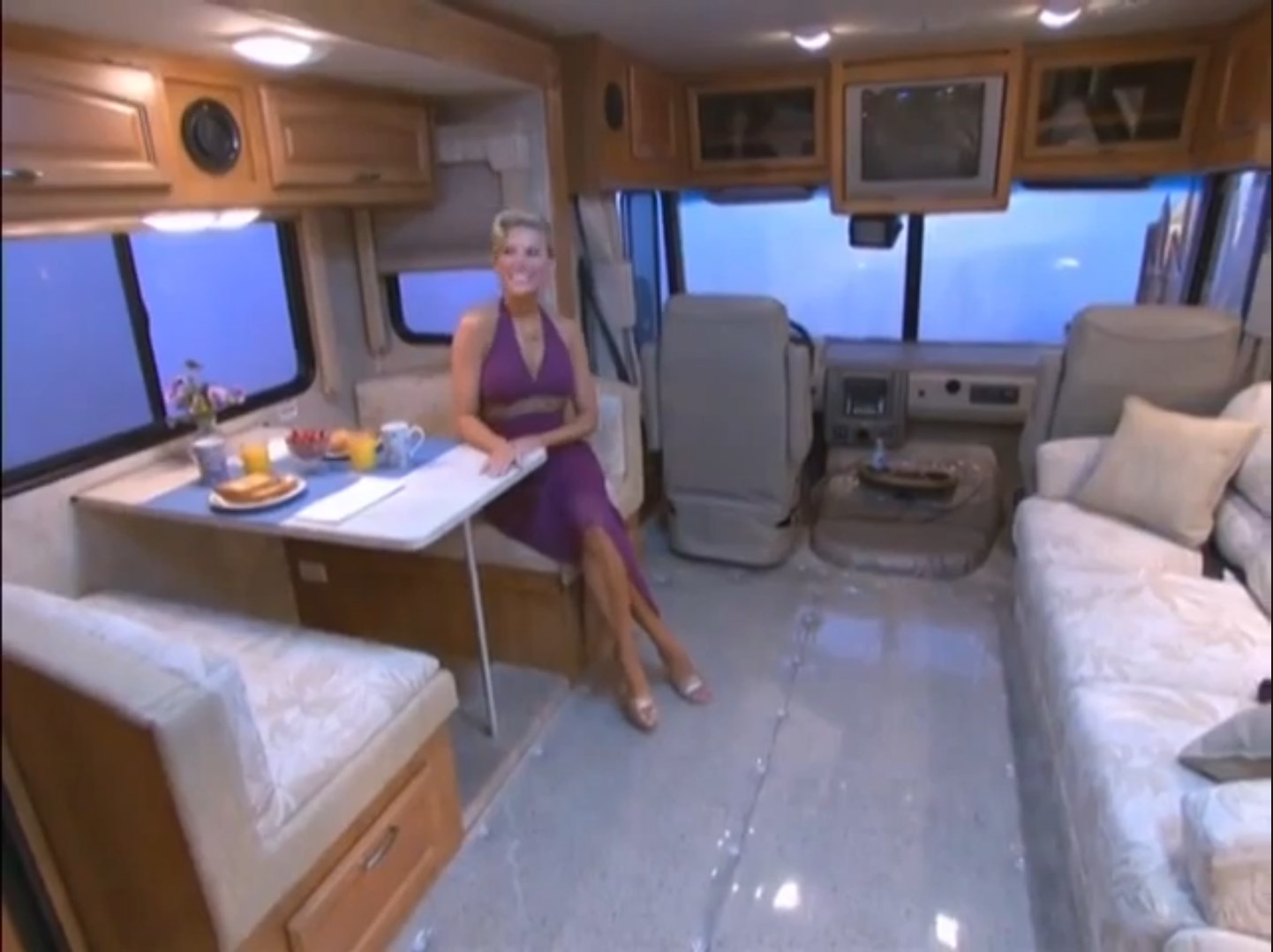 A model sits at a dining table in a luxury motorhome, enjoying breakfast with a view out the windows.