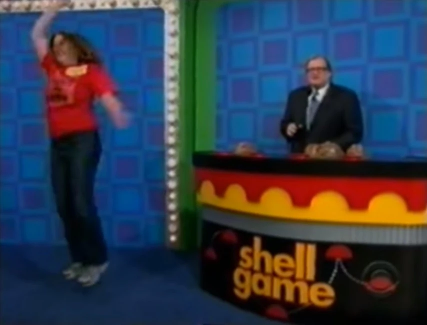 A contestant enthusiastically jumps while participating in the Shell Game on The Price is Right, with a host standing beside a brightly colored game setup.