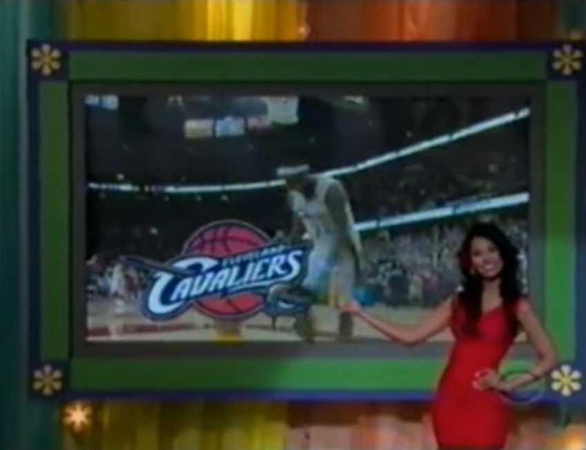 A woman in a red dress smiles and points to a large screen displaying the logo of the Cleveland Cavaliers basketball team, with a player in action on the court behind her.