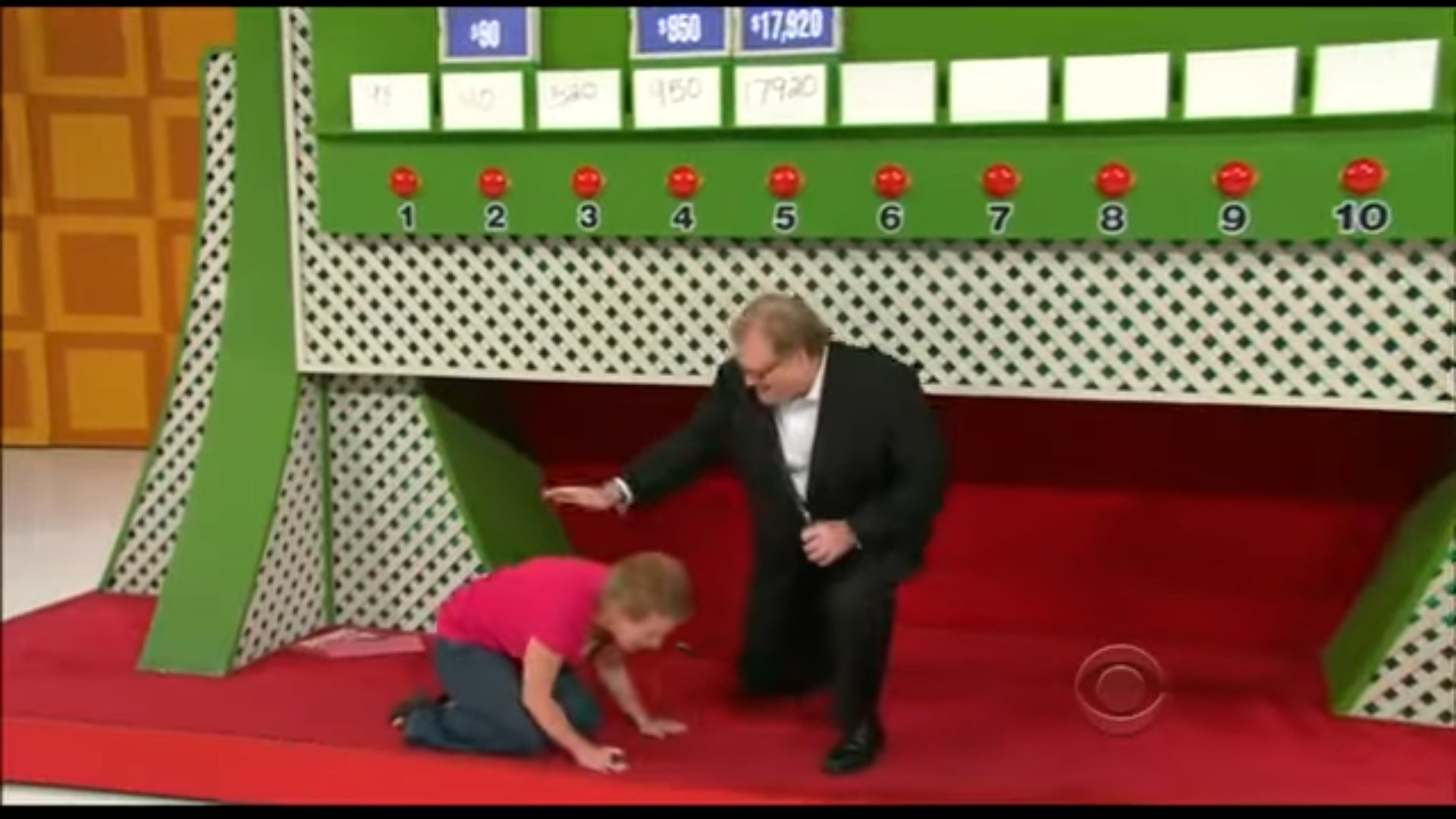 A host kneels beside a contestant, who is on the floor looking for a hidden item during the game Ten Chances on The Price is Right set.