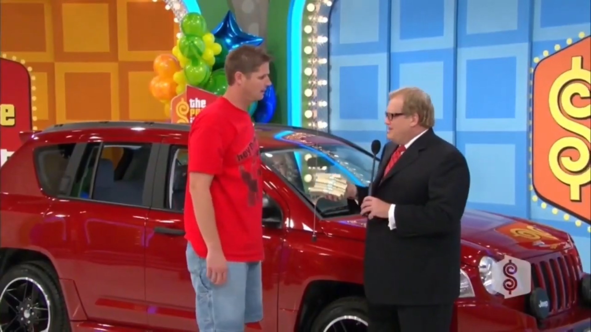 A contestant on a game show stands next to a red car, while the Drew Carey them a stack of ,000 in a special playing of Lucky Seven.