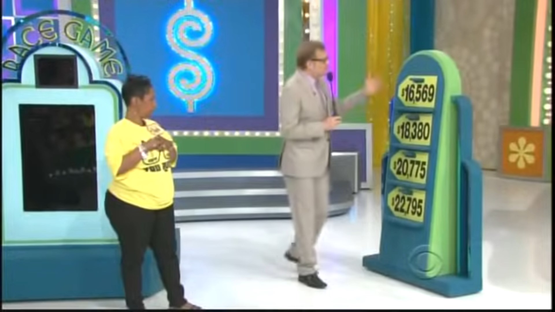 A contestant in a yellow shirt stands next to a game host, who gestures towards a board displaying four price tags in a colorful game show setting.