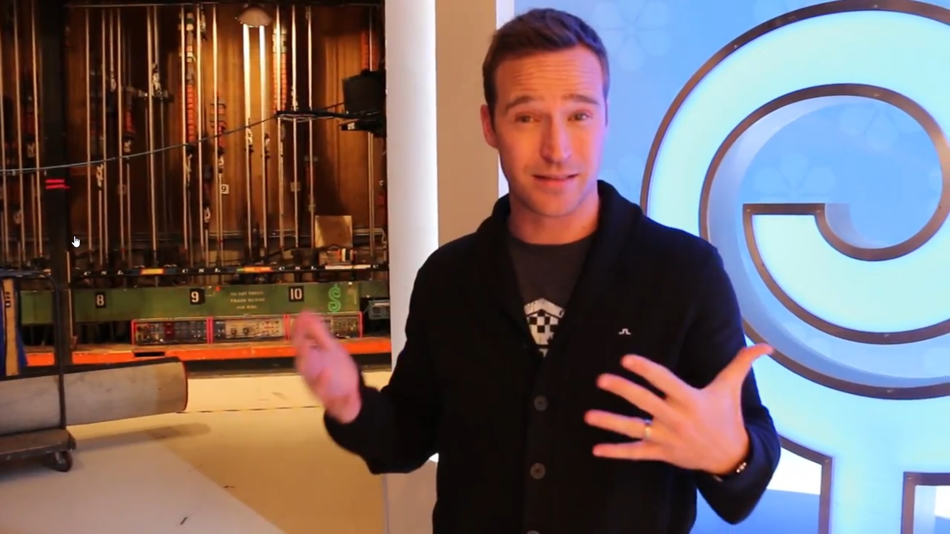 A host explaining the setup of a card game show with colorful studio lights and equipment in the background.