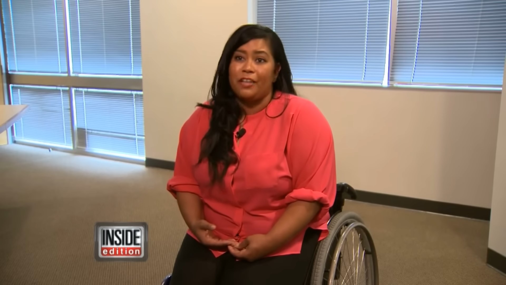 A woman in a pink blouse is sitting in a wheelchair, speaking during an interview in a well-lit room.
