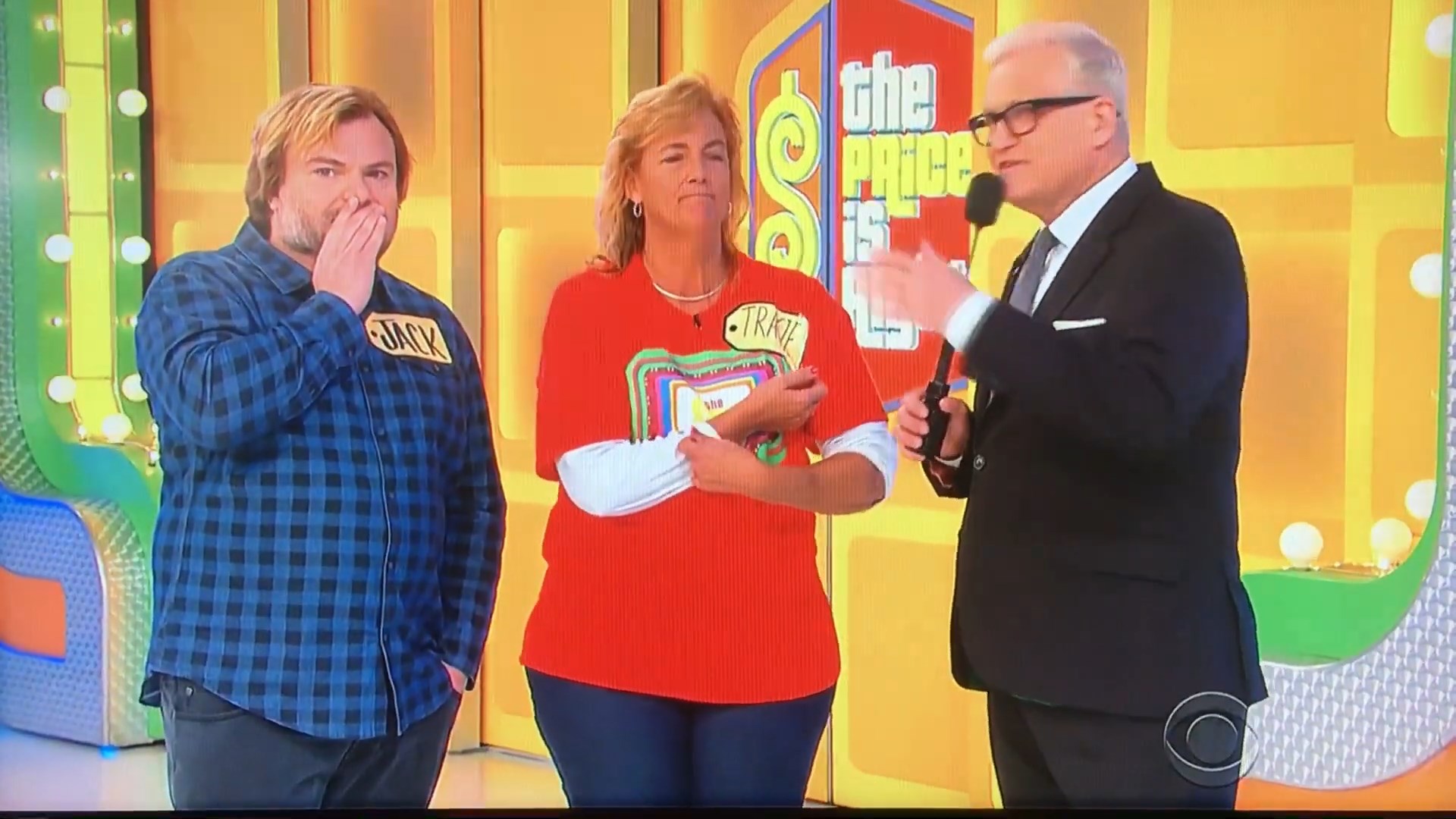 A contestant named Tracie listens intently while Jack Black, who is next to her, contemplates his guess during a game segment on The Price Is Right. Drew Carey is speaking to them, with a colorful backdrop behind them.