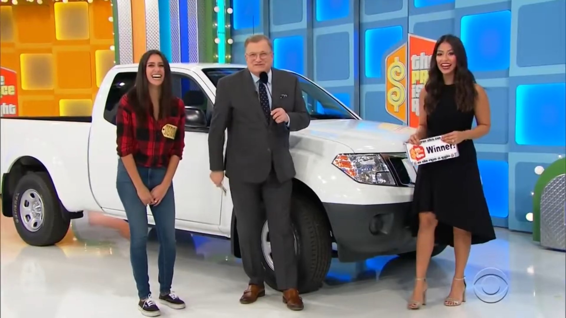 A jovial contestant smiles while standing next to a white pickup truck on stage, alongside a host and another woman holding a 'Winner' sign.