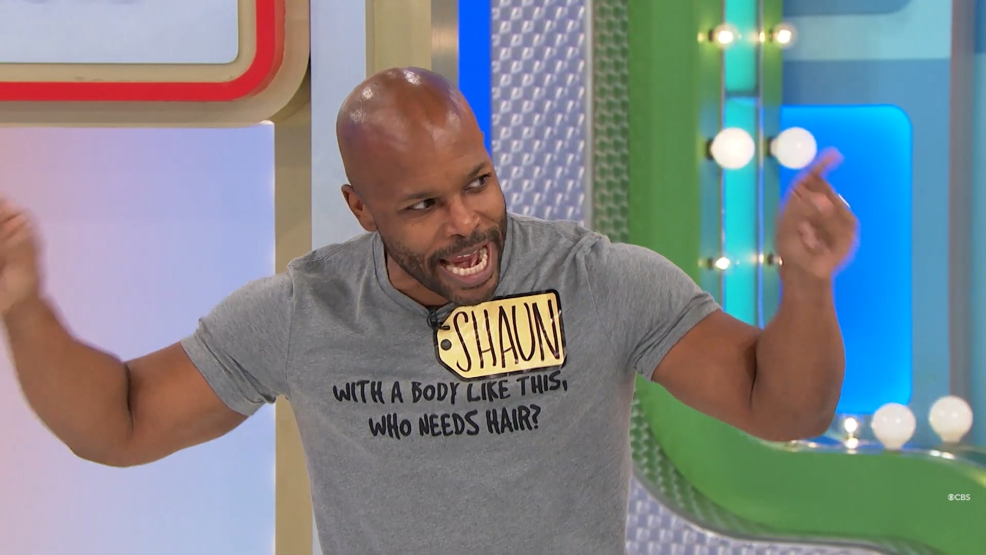 A contestant named Shaun enthusiastically participates in a game show, wearing a humorous T-shirt that reads, 'WITH A BODY LIKE THIS, WHO NEEDS HAIR?'