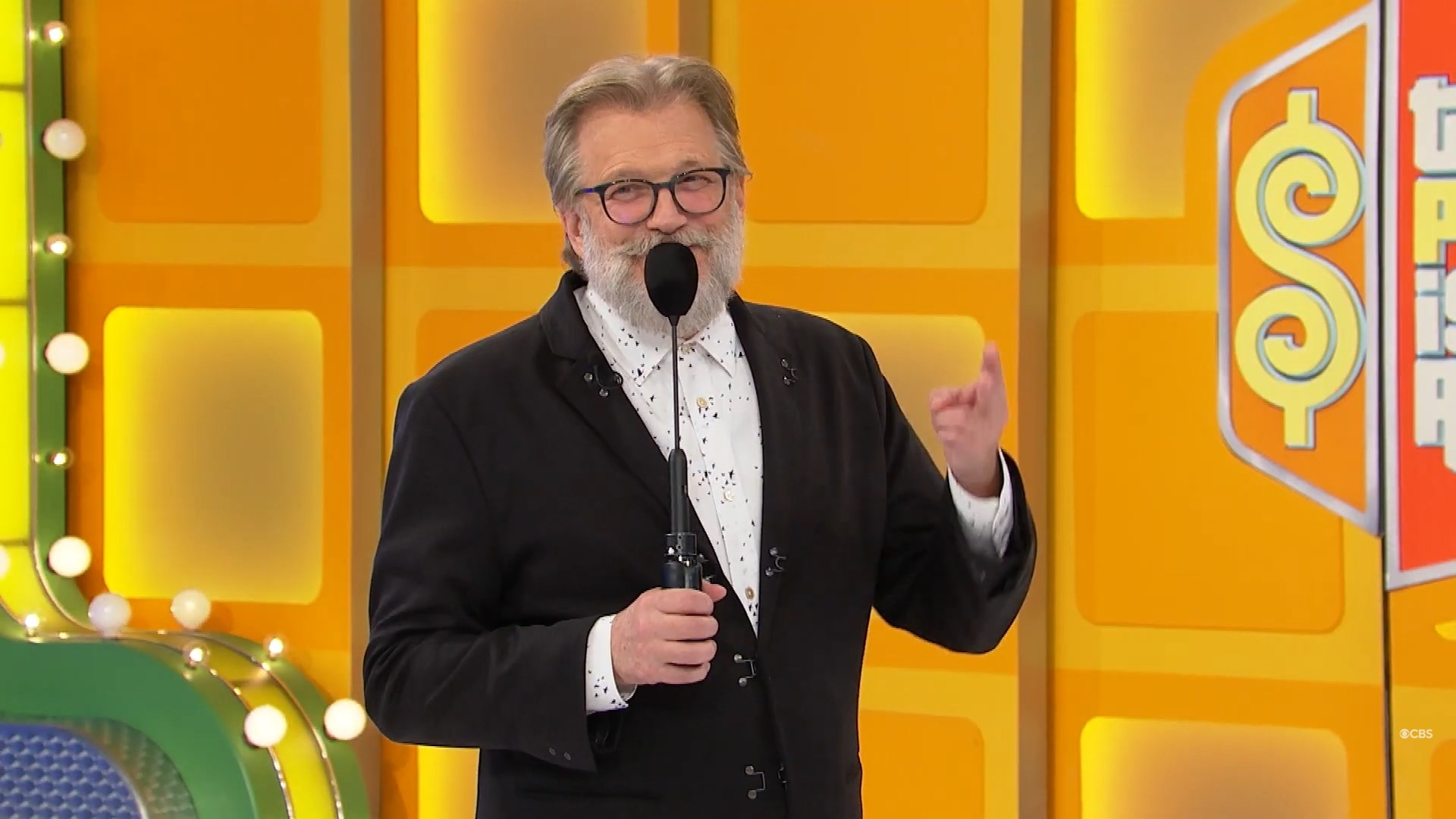 Drew Carey with a beard and glasses is speaking into a microphone on a colorful game show set.