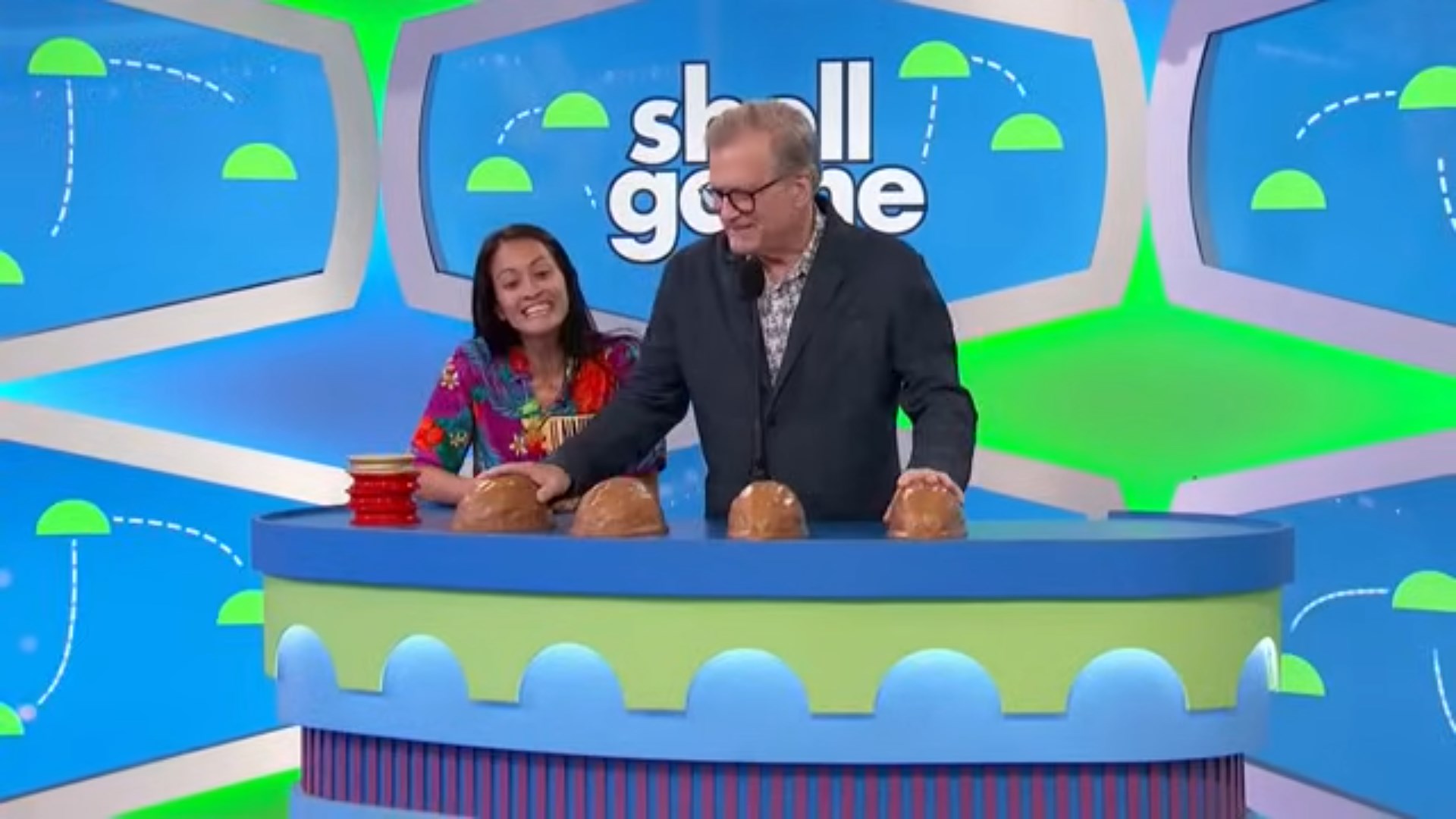 A male host and a female contestant are playing the Shell Game, standing behind a table with three shells on it. The set features a colorful background with gaming graphics.