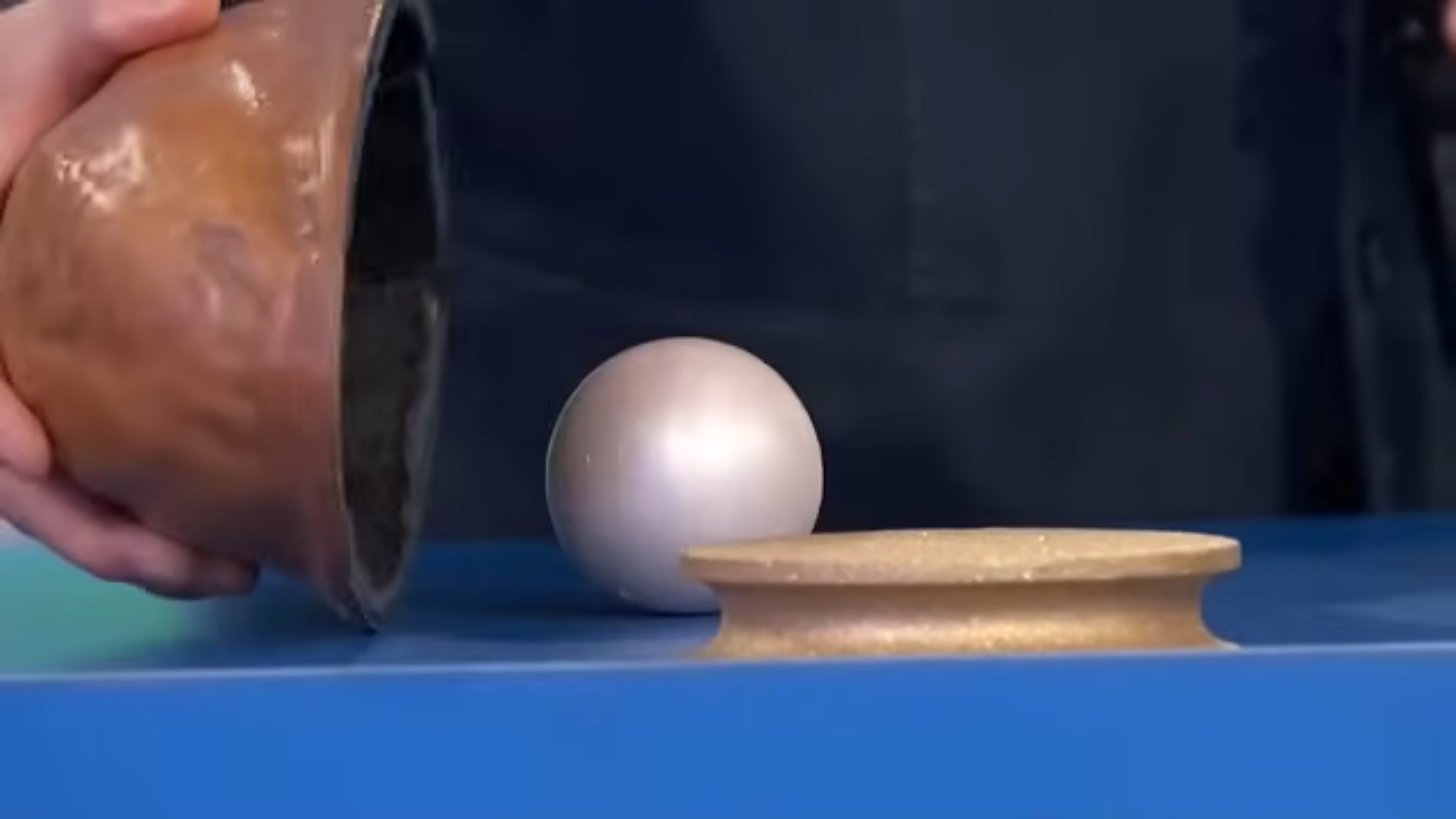 A hand holding a shell is revealed, while a shiny ball sits on a table beside it.
