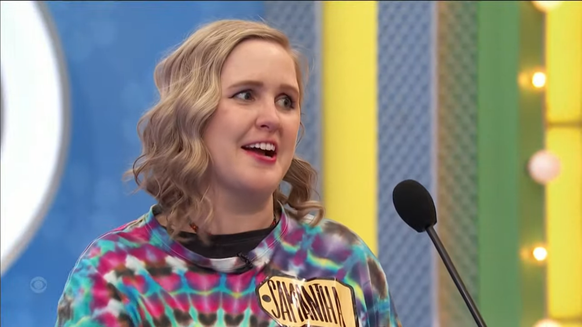 A contestant named Samantha smiles nervously while competing on a game show, wearing a colorful tie-dye shirt and holding a name tag.