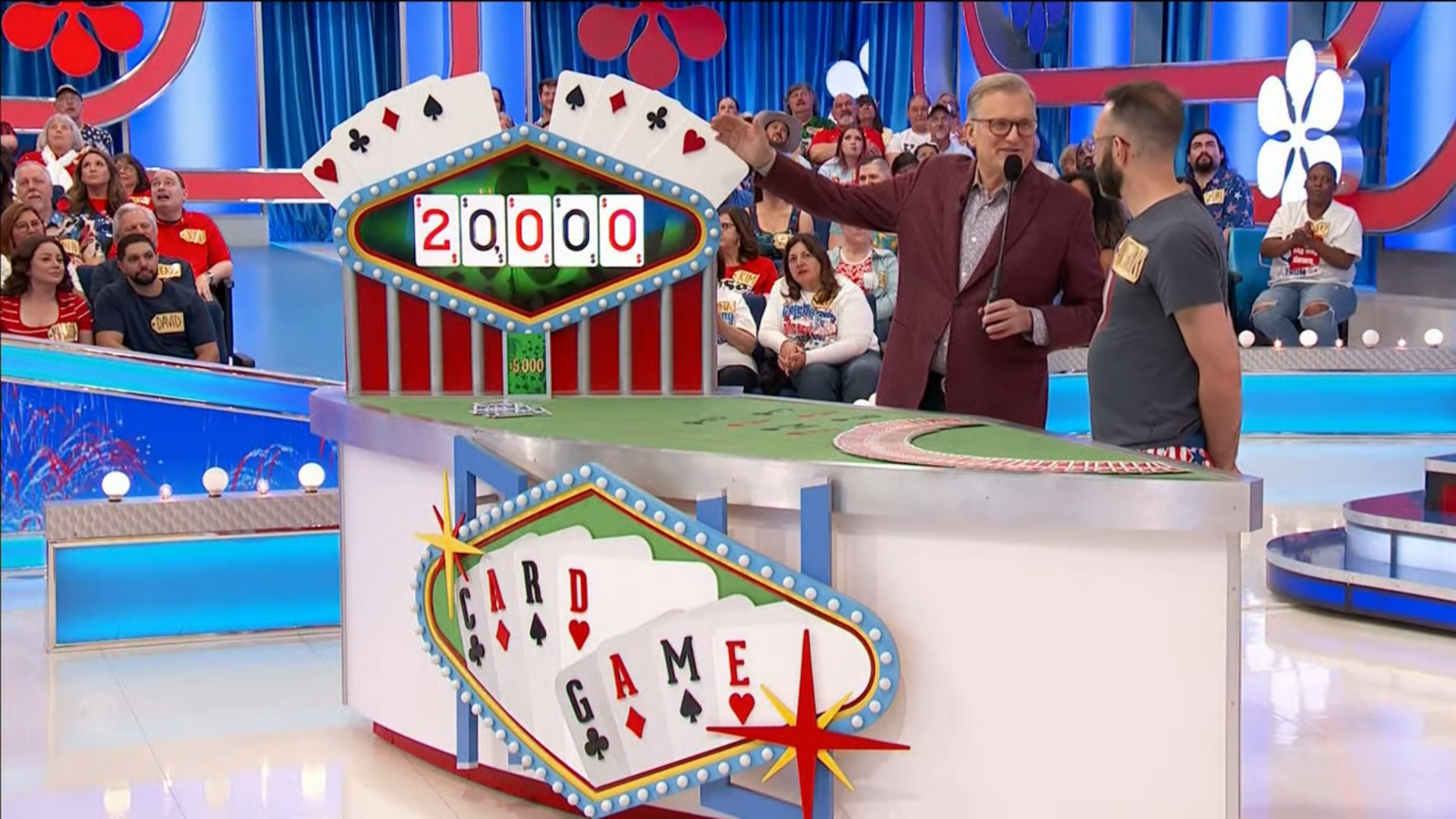 A game show host presents the 'Card Game' with a scoreboard indicating a starting bid of ,000, while a contestant stands beside him at the game table, amidst an enthusiastic audience.