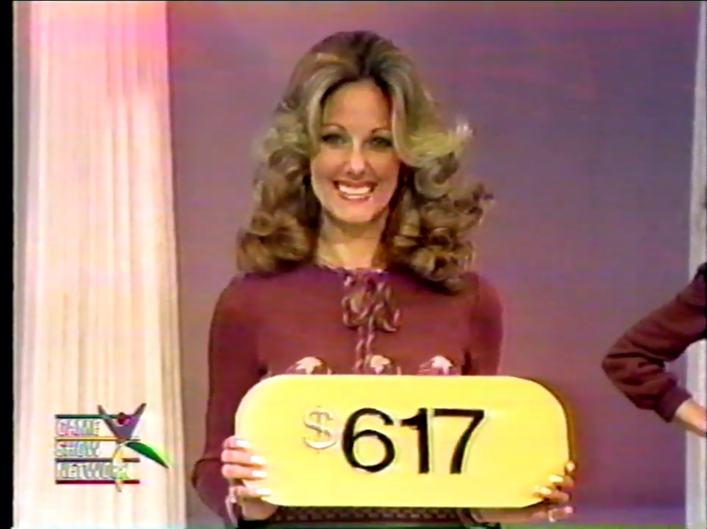 A model smiles while holding a yellow price tag displaying 7 on a game show set.