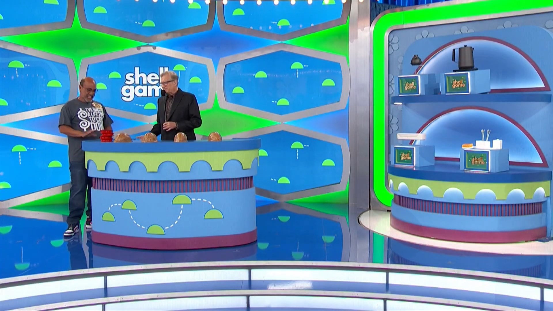 A game show host stands next to a contestant at the Shell Game setup, featuring colorful backgrounds and three shells on a table.
