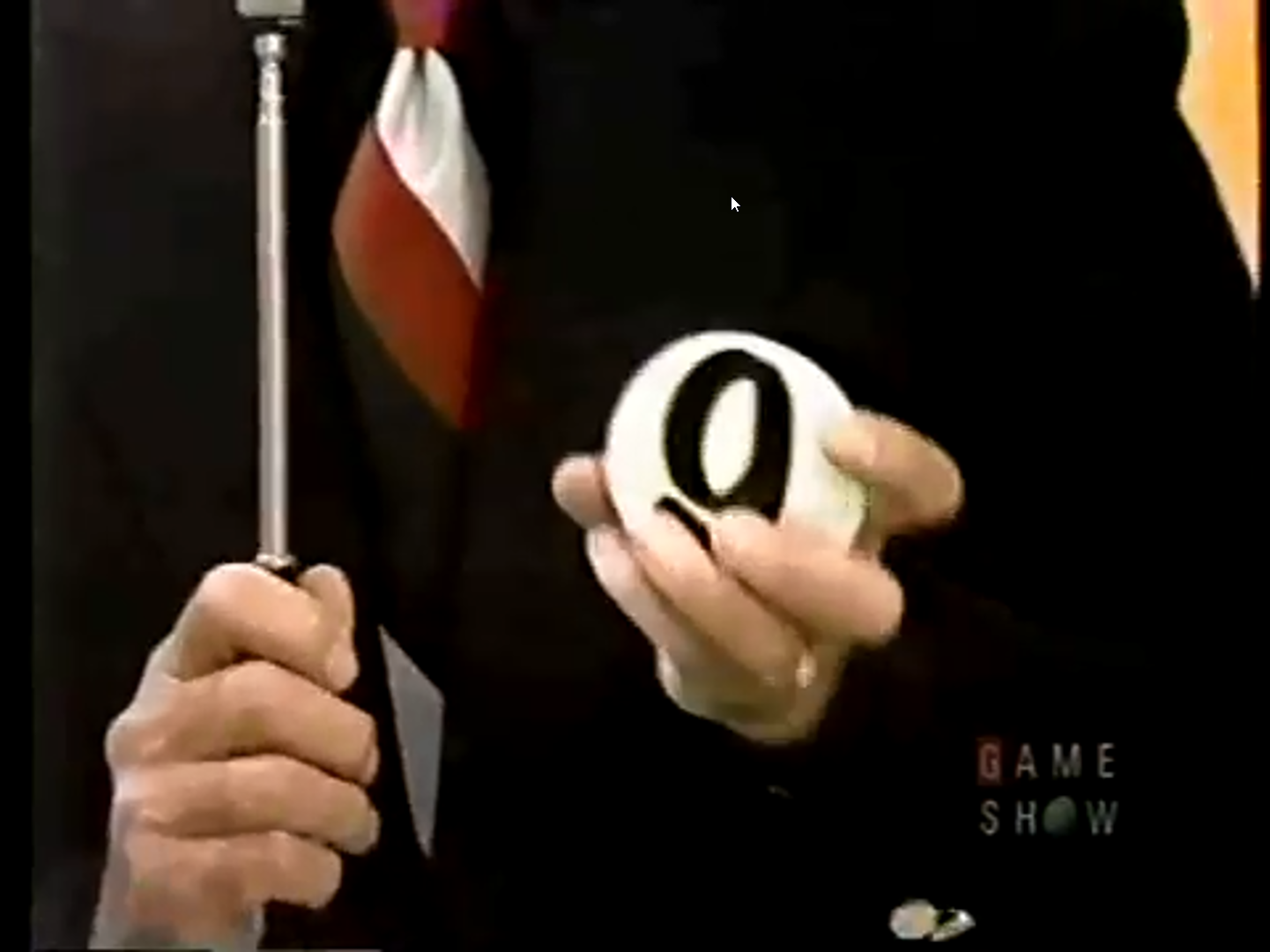 A game show host holds a white ball with the number '9' printed on it, while gripping a microphone with a colorful striped tie visible.