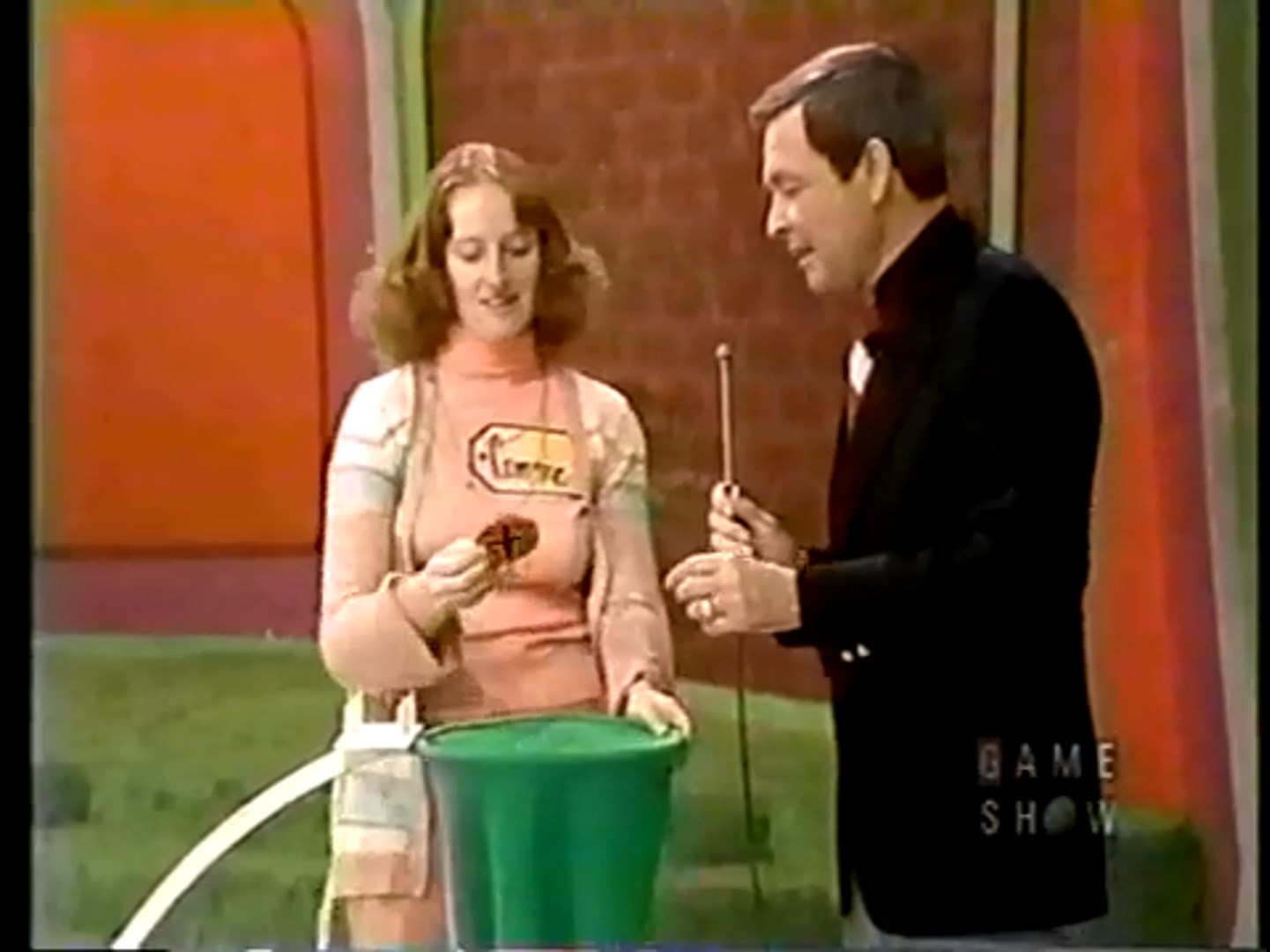 A contestant smiling while holding a chip drawn from a green bag, with a game show host standing beside her, in a colorful studio backdrop.