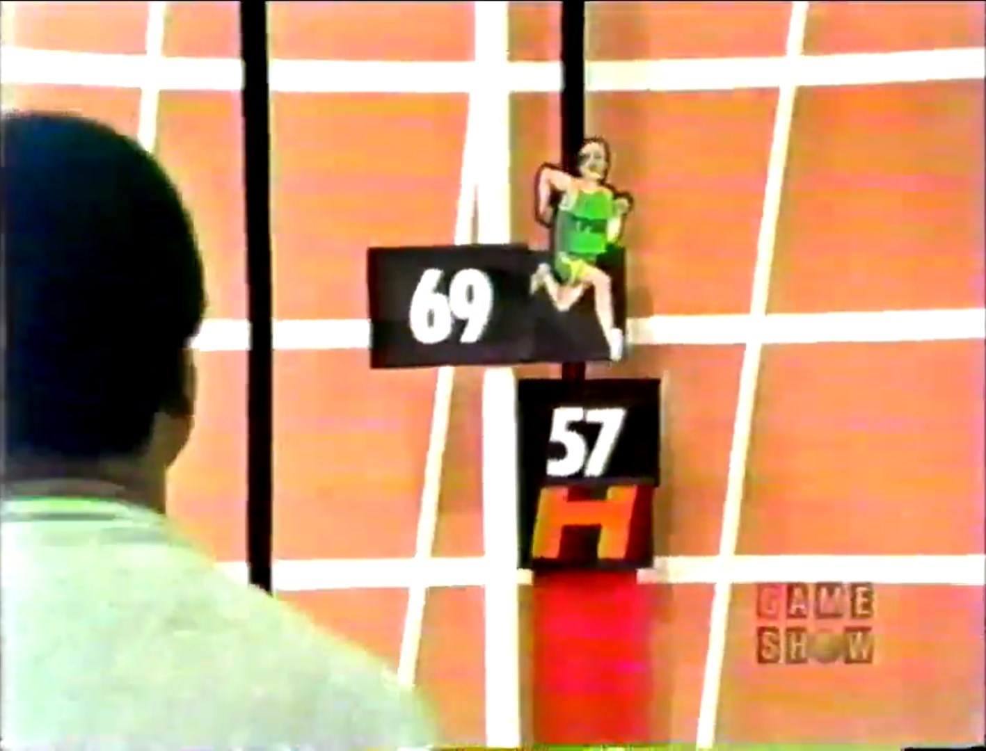 A contestant watches as a cartoon runner jumps over hurdles labeled with grocery prices on a game show set.