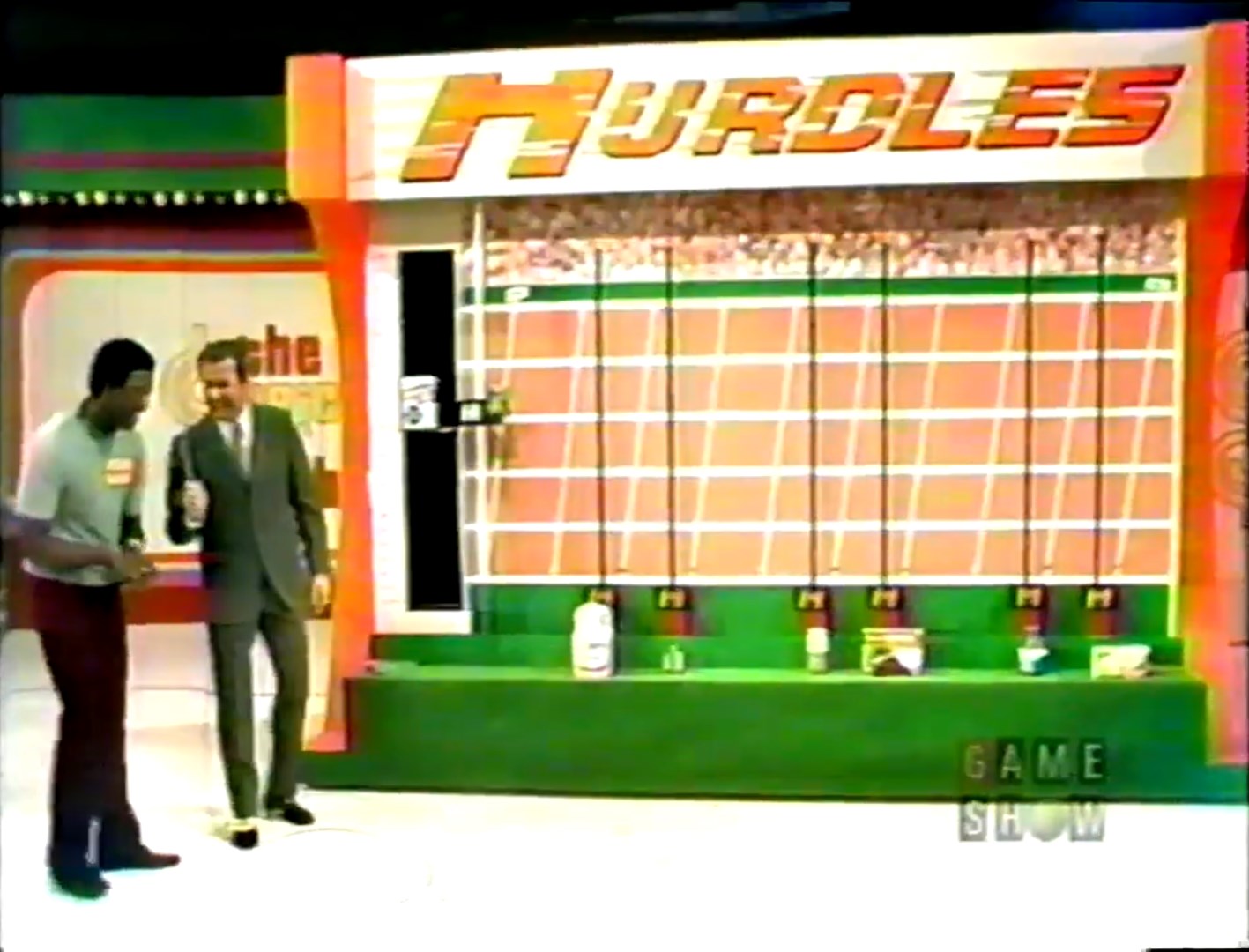 A contestant and host stand beside the Hurdles game setup on The Price is Right, featuring a large board with three hurdles and various grocery products displayed below.
