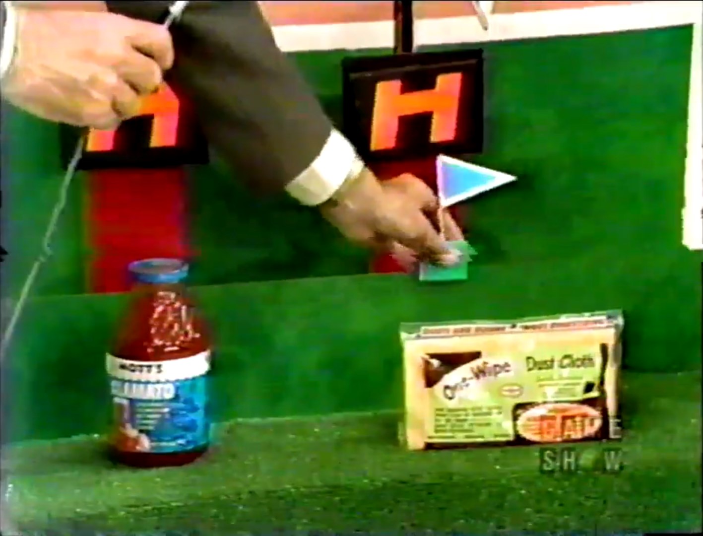 A hand holds a flag marker near a game board featuring a bottle of Mott's tomato cocktail juice and a package of One-Wipe dust cloths, on a lush green surface.