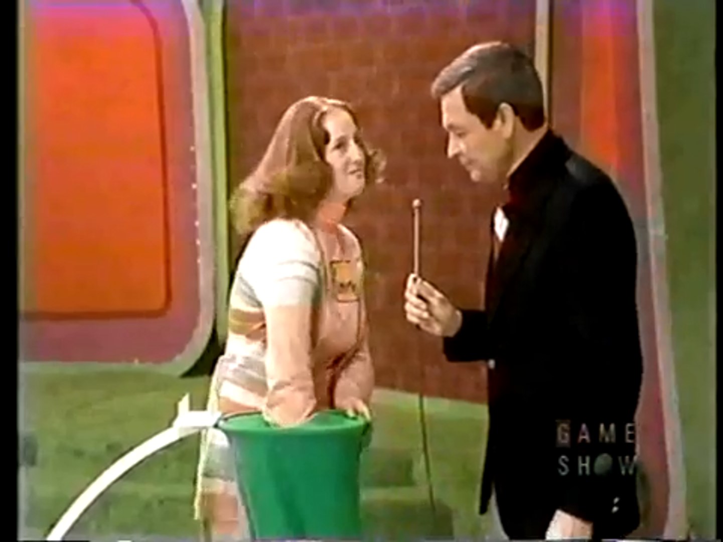 A contestant smiles while interacting with the host on a game show stage, featuring a green container and colorful backdrop.
