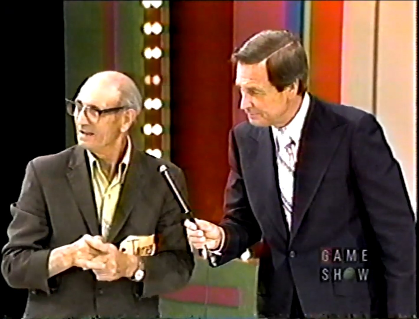 A contestant with glasses and a suit appears on The Price is Right stage, speaking to Bob Barker who is holding a microphone, with colorful set lights in the background.