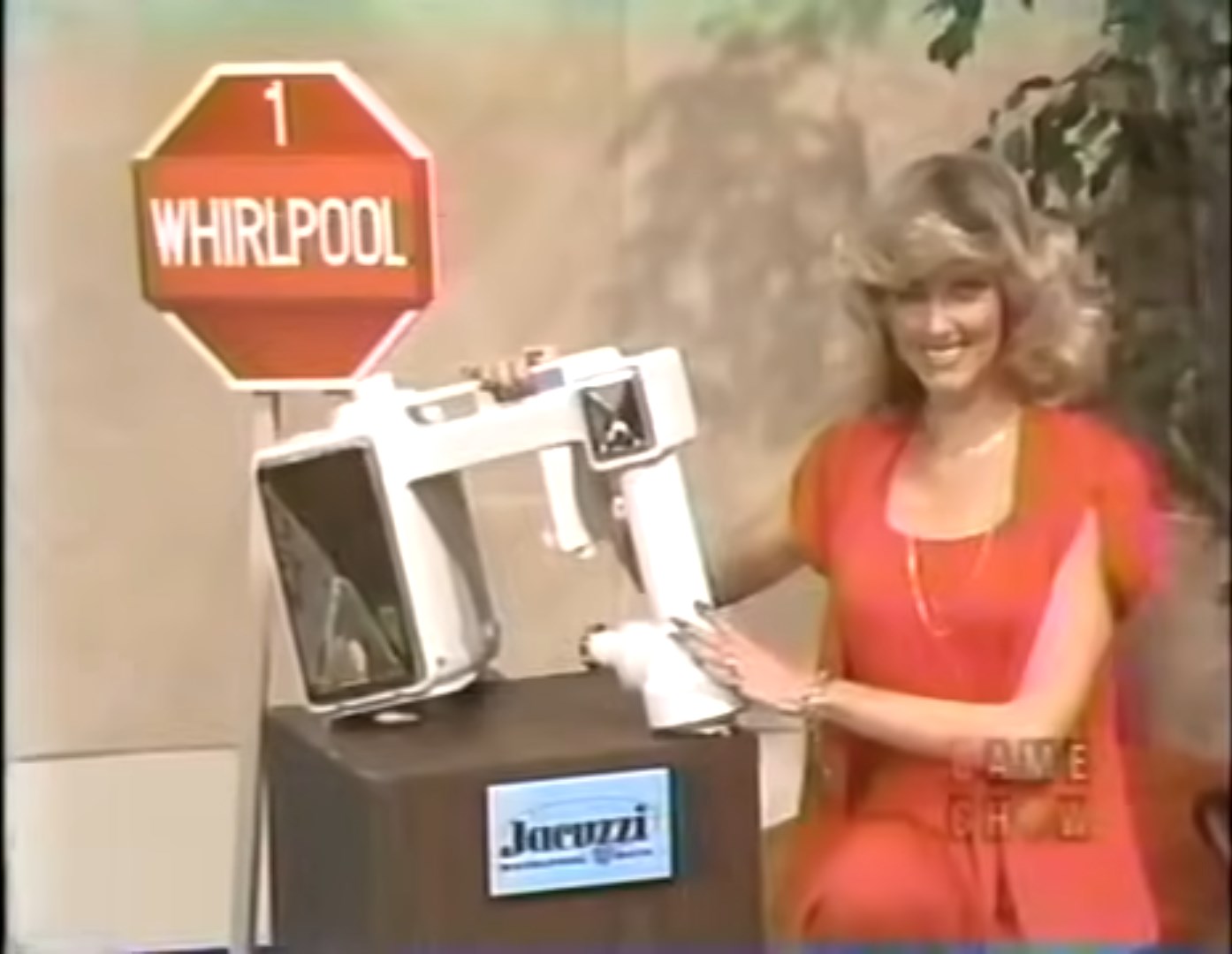 A woman in a red outfit stands next to a sign that says '1 WHIRLPOOL,' holding a small whirlpool bath unit on a podium labeled 'Jacuzzi.'