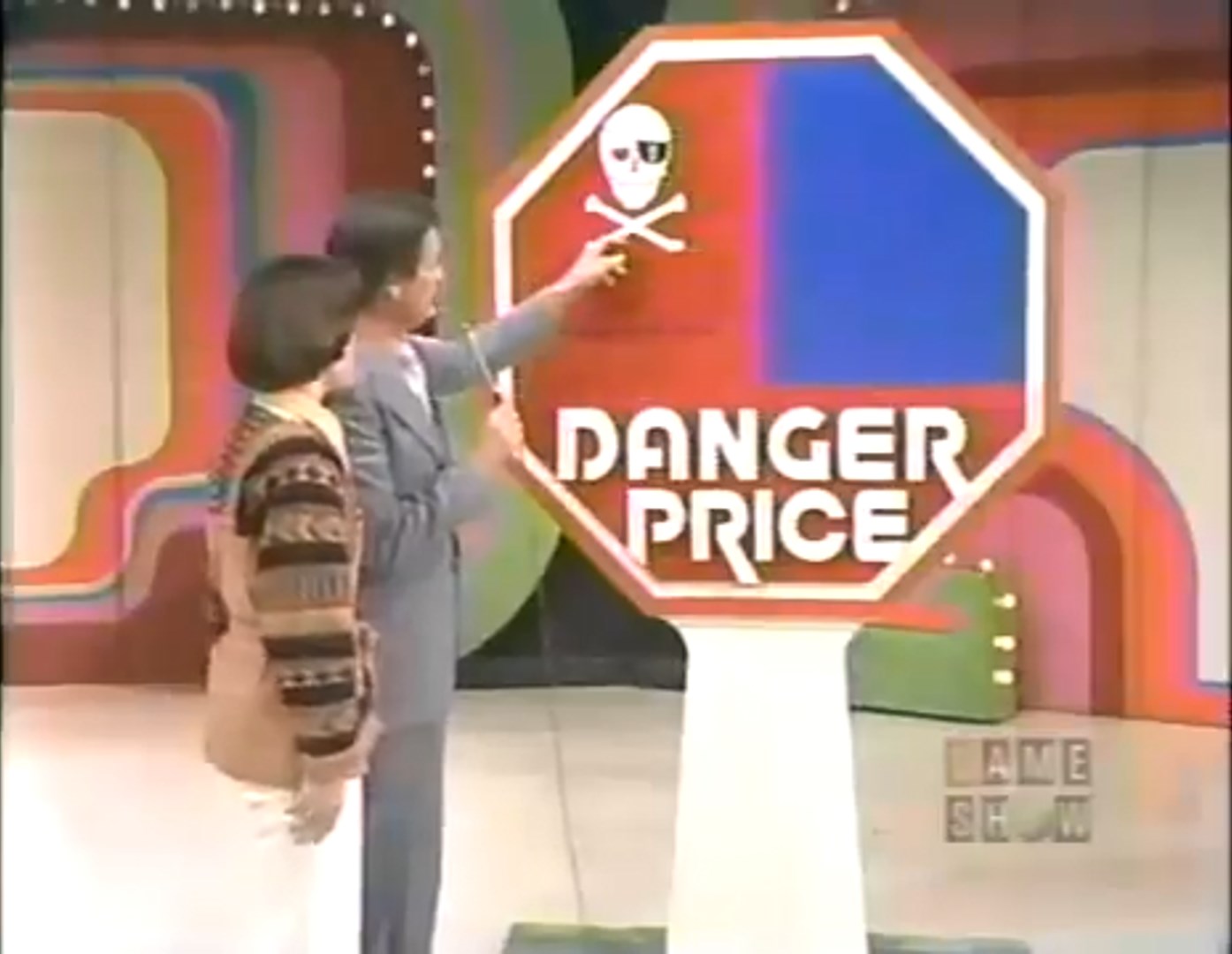 A contestant and host point to the 'Danger Price' sign on a game show set, featuring a skull and crossbones symbol.