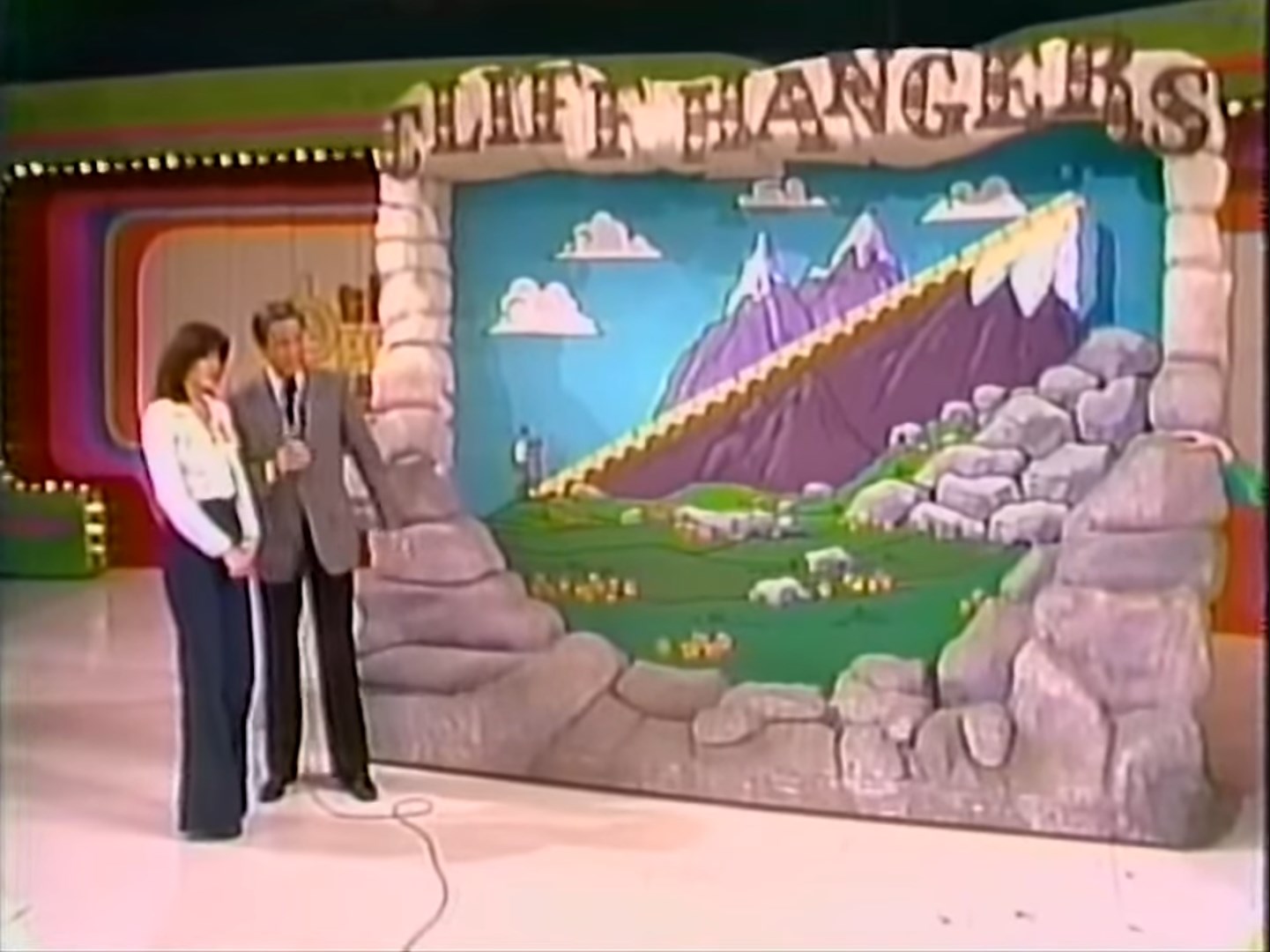 A host and contestant stand beside the animated game set for 'Cliff Hangers,' featuring a mountain scenery and a staircase illustration.