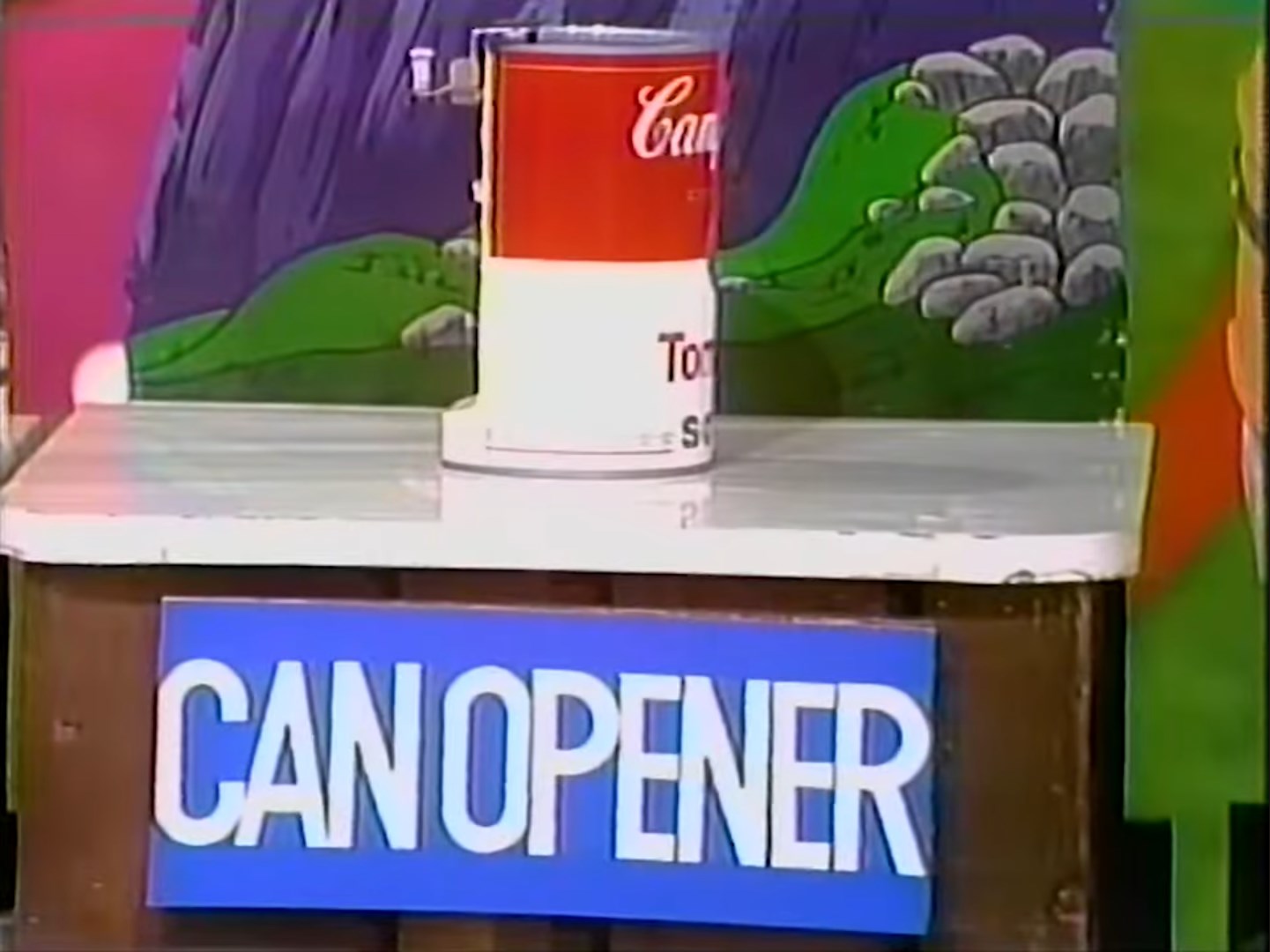 A can opener displayed on a game show set with a colorful mountain backdrop.