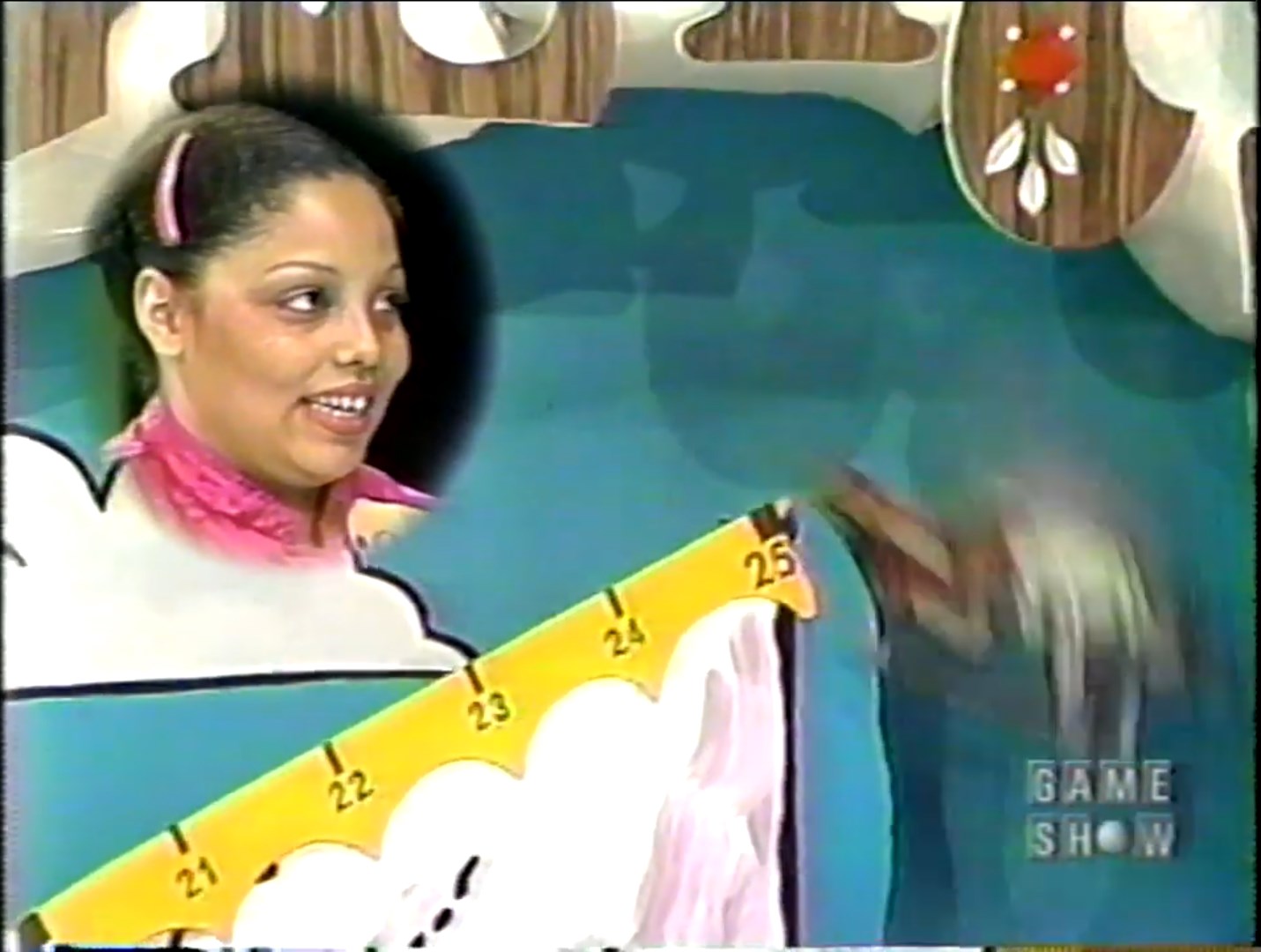 A contestant smiles while playing Cliff Hangers, with a colorful game board displaying a mountain and a scale marked 0 to 25.