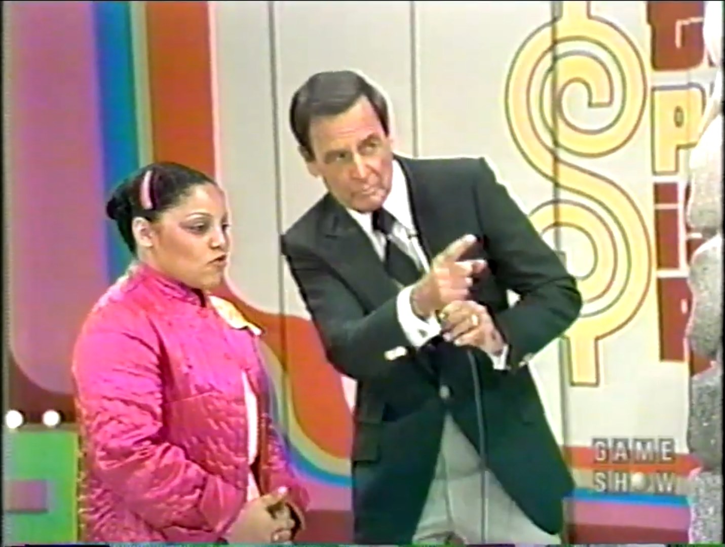A contestant in a pink jacket looks serious while standing next to a host in a tuxedo, as they prepare for a segment on The Price is Right game show.