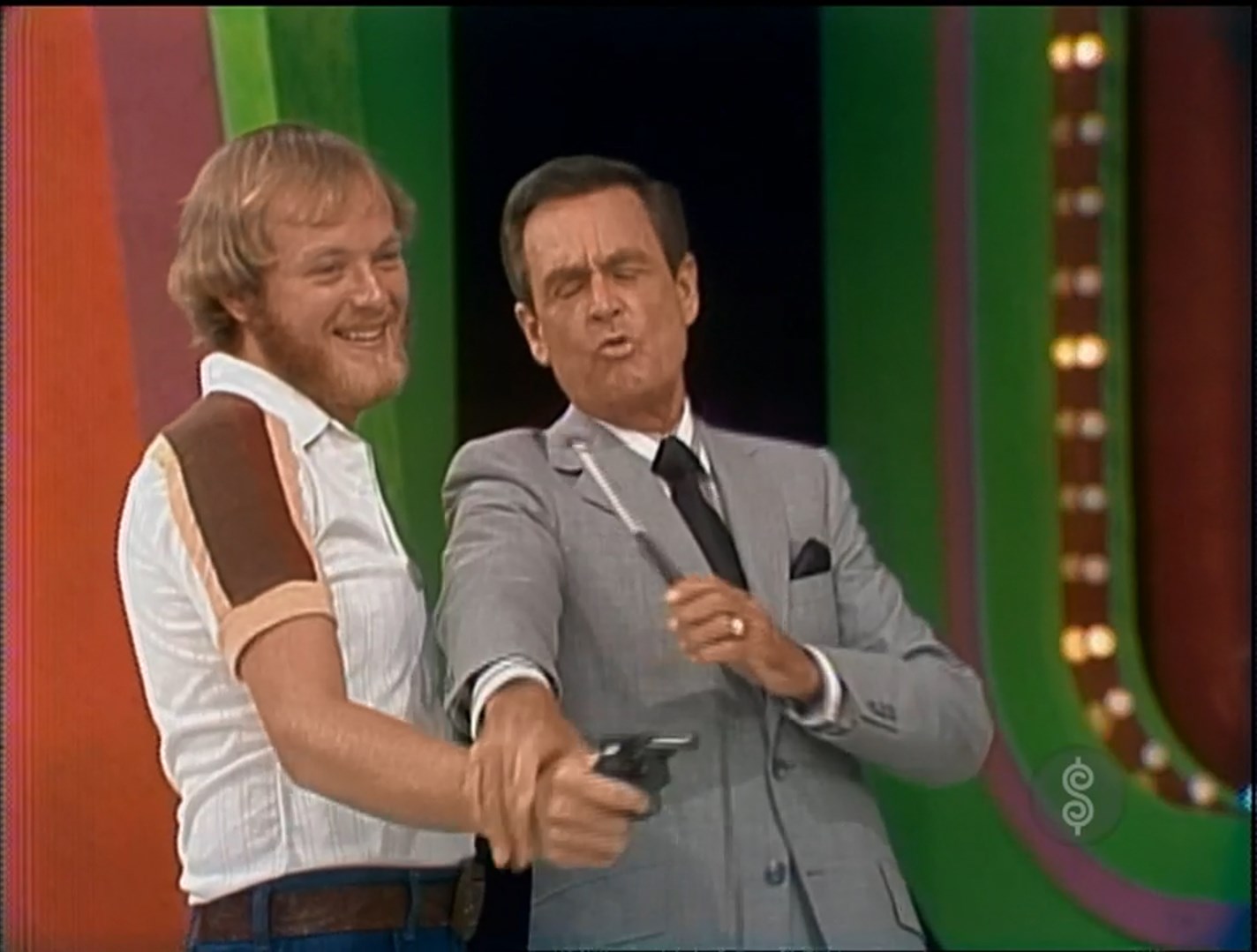 A contestant and a host on a game show are playfully aiming a toy gun, with the contestant smiling and the host making a comedic face.