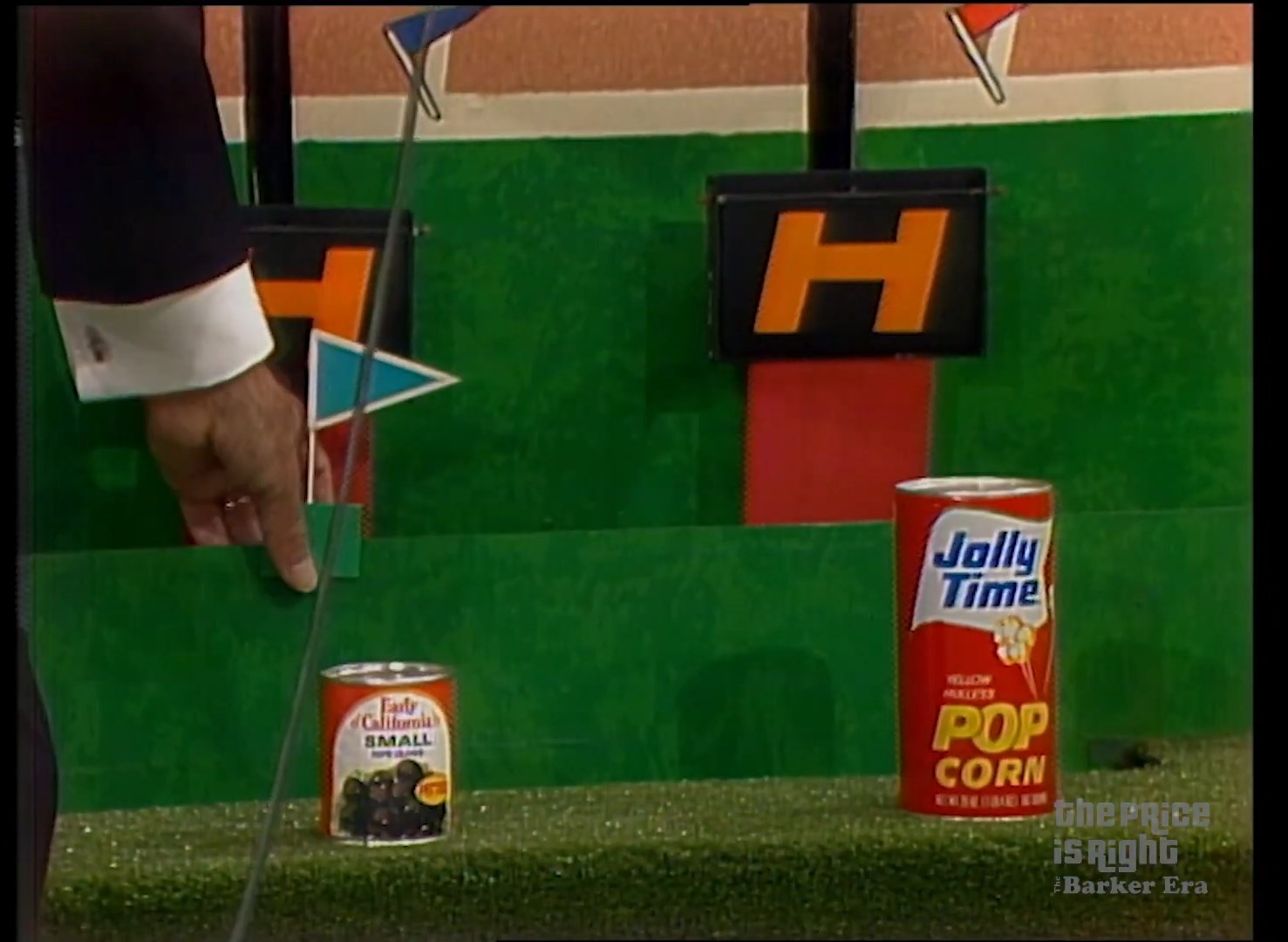 A close-up of a hand pointing at two grocery products on a game show set. One is a can of 'Early California's Small' olives, and the other is a can of 'Jolly Time' popcorn. The backdrop features a colorful design with the letter 'H' for the game Hurdles.