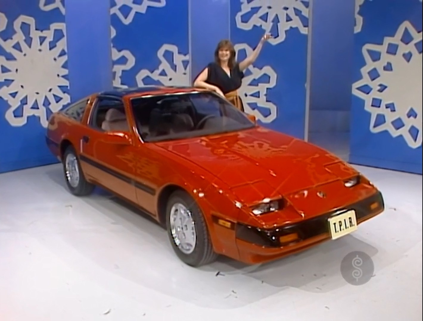 A model stands next to a bright orange 1986 Nissan 300ZX sports car on a game show set, pointing excitedly at the car.