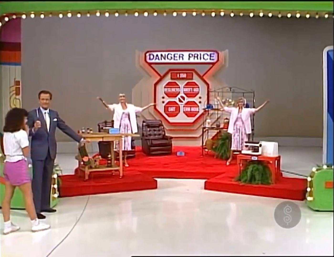 A game show set featuring the 'Danger Price' game with a host and contestants. The stage displays the game's title alongside various prizes, including furniture and a sewing machine.
