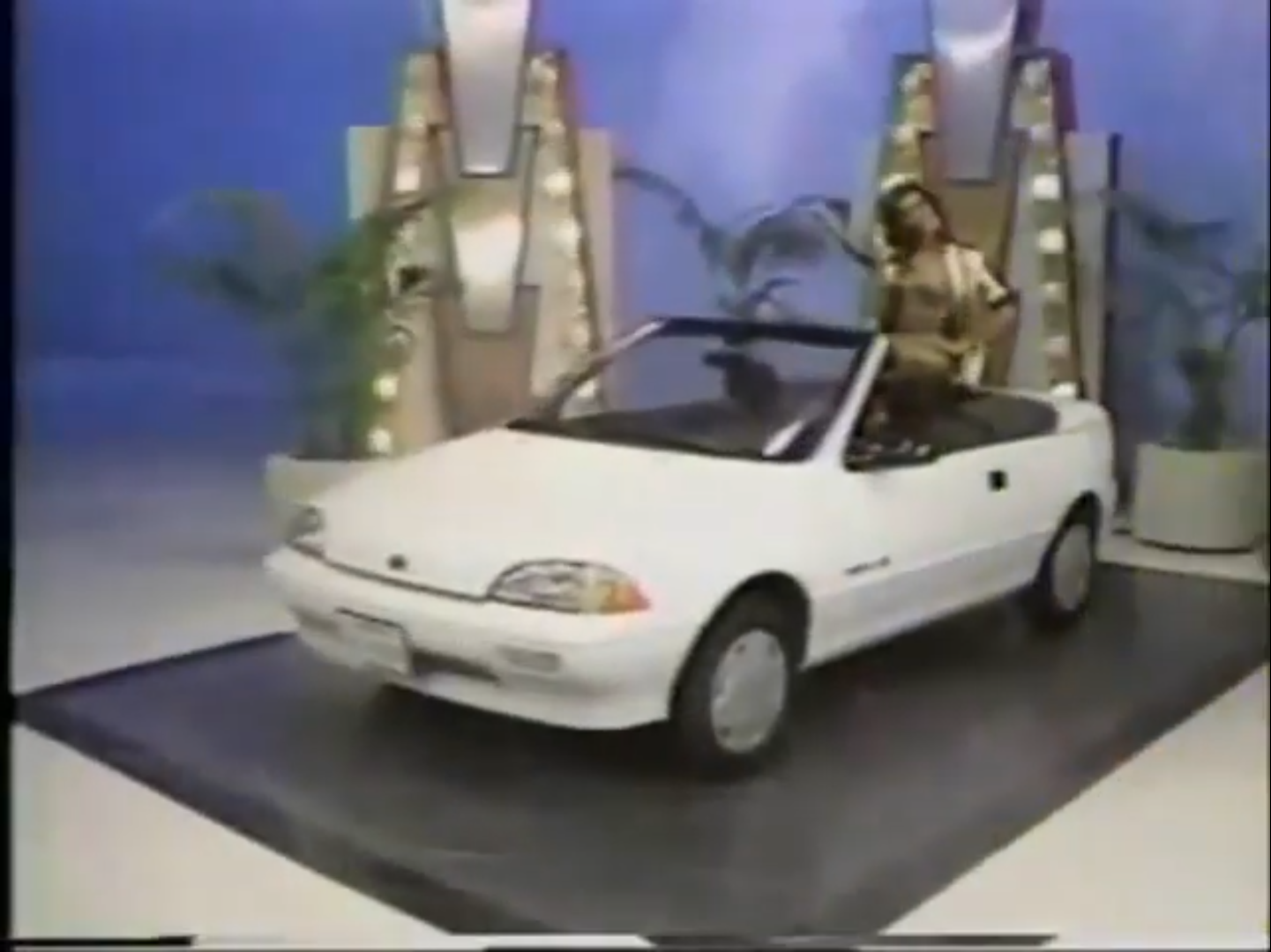 A model poses in a white convertible car on a stage with bright lights and colorful decorations.