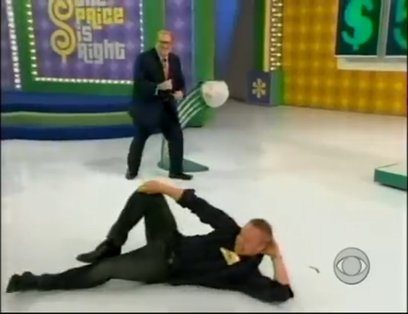 A contestant lies on the floor of a game show set while Drew Carey stands nearby, creating a playful and humorous moment.