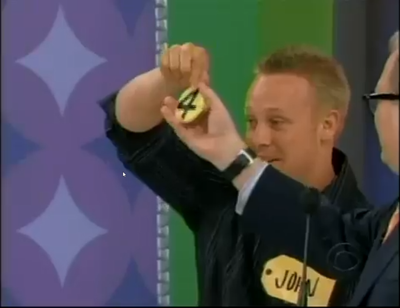 A contestant named John holds up a yellow chip displaying the number 4, while a host watches nearby.