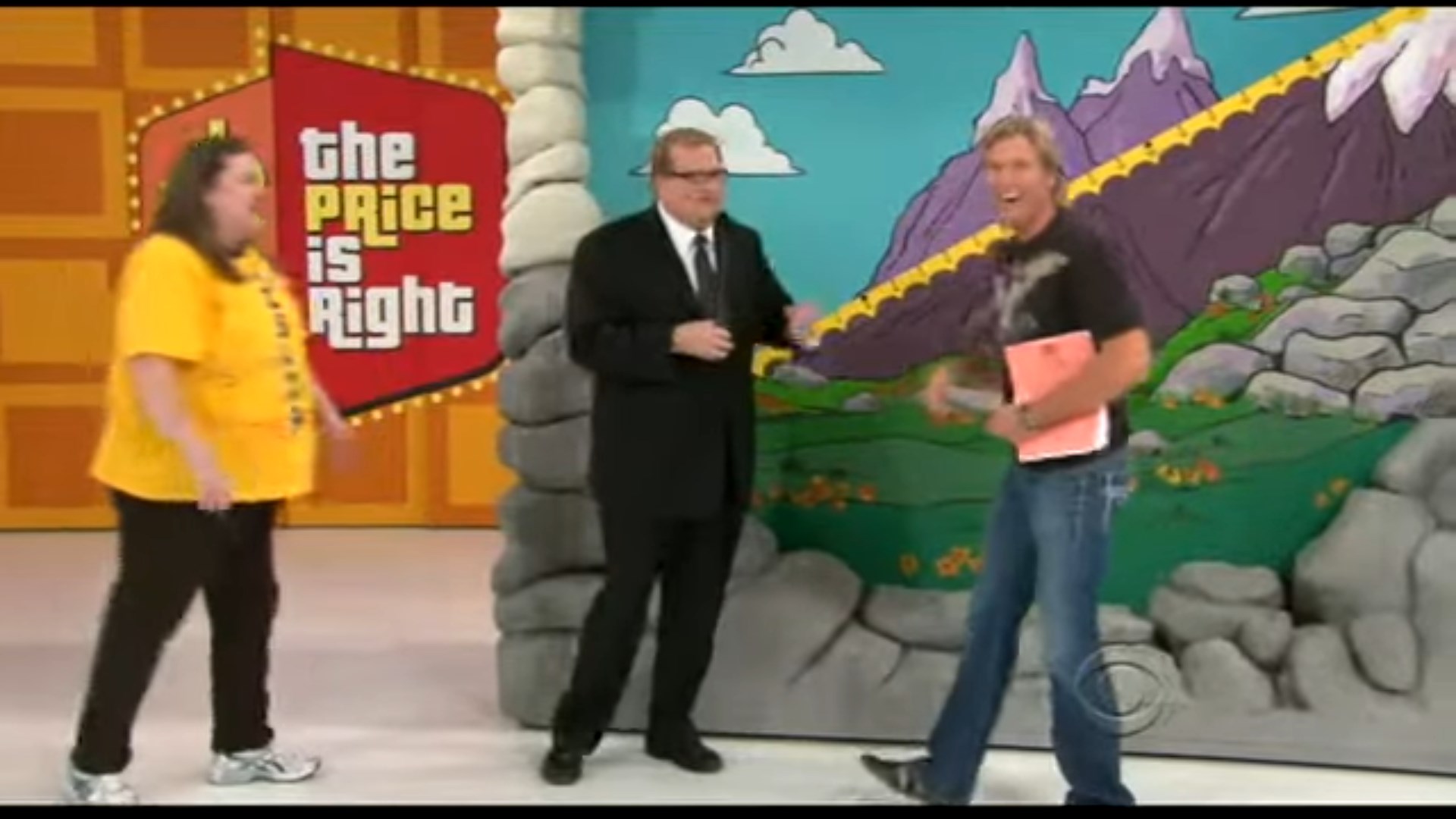 Jack Wagner, holding a script, interacts with contestant and host Drew Carey near the Cliff Hangers game set during an episode of The Price is Right, featuring a colorful mountain backdrop.