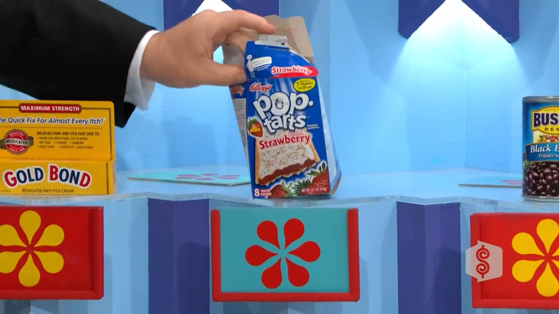 A hand is holding a box of Kellogg's Strawberry Pop-Tarts, with a Gold Bond Anti-itch cream box in the foreground on a brightly colored game show set.