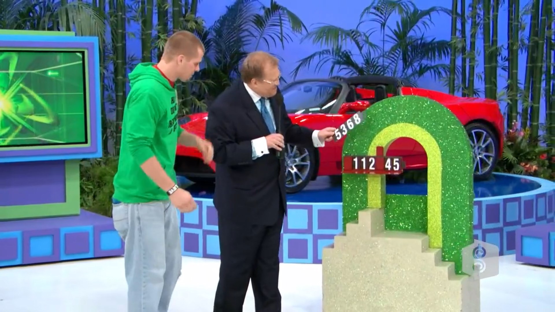 A contestant and host interact while revealing digits for the Golden Road game, with a luxury red Tesla Roadster in the background.