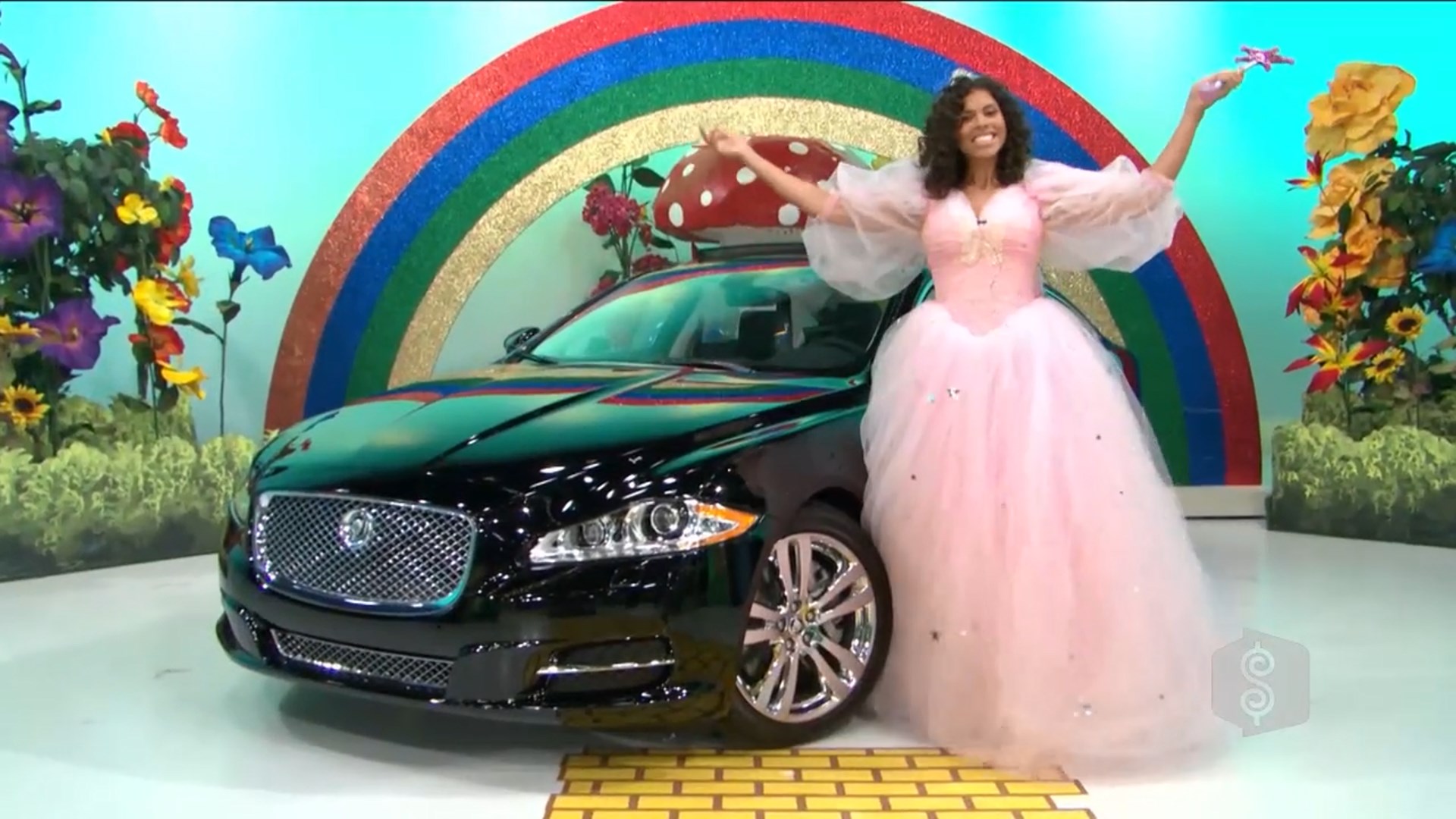 A model in a pink ball gown stands next to a black luxury car, smiling and gesturing joyfully in front of a colorful, rainbow-themed backdrop adorned with flowers.
